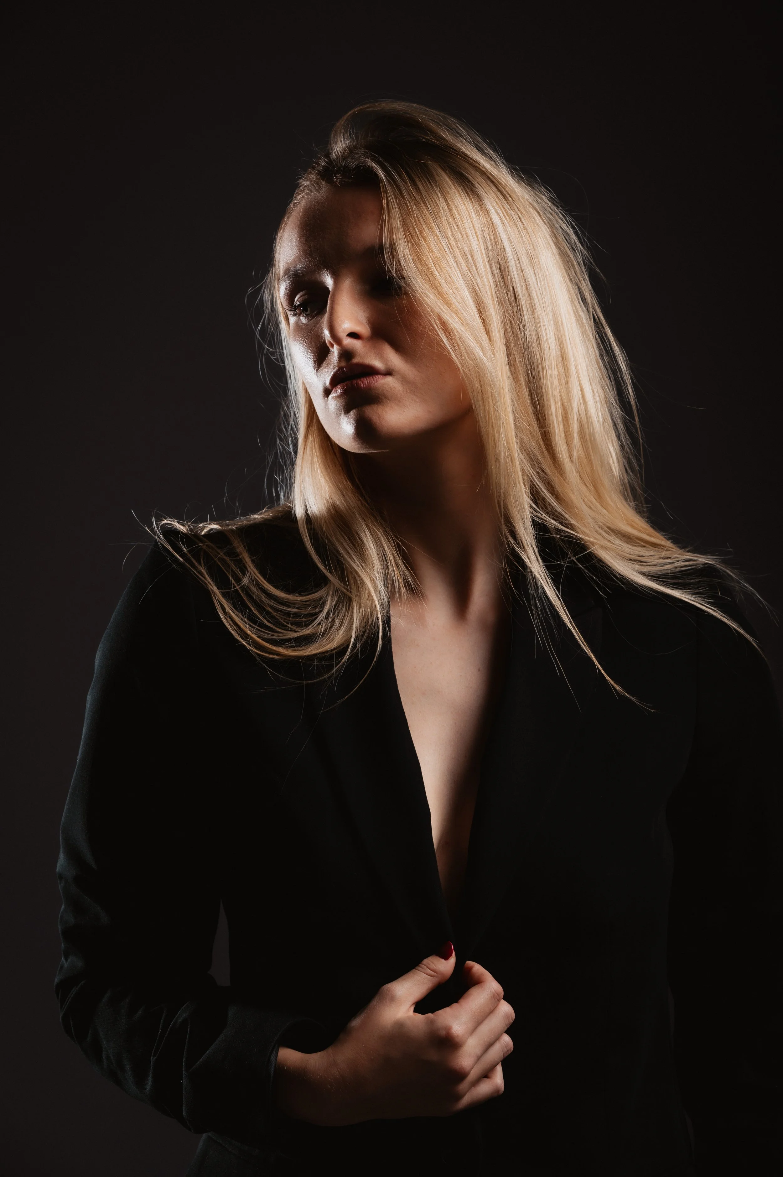 Woman with long blonde hair wearing a black jacket, in a dark, moody setting.