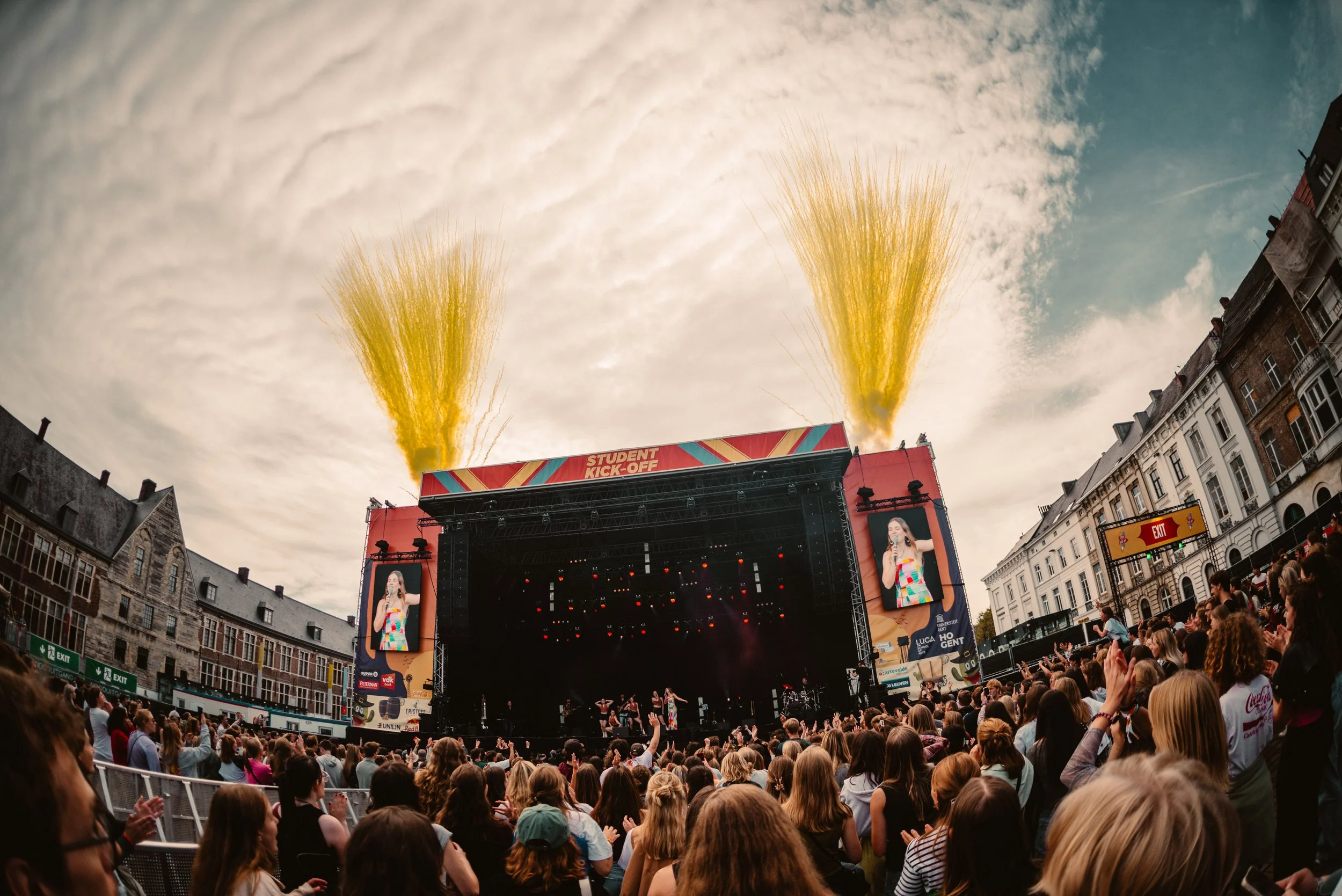 Outdoor concert with large crowd in front of a stage labeled 'Student Kick-Off'; two large screens display a performer; yellow smoke or confetti is emitting from the stage.
