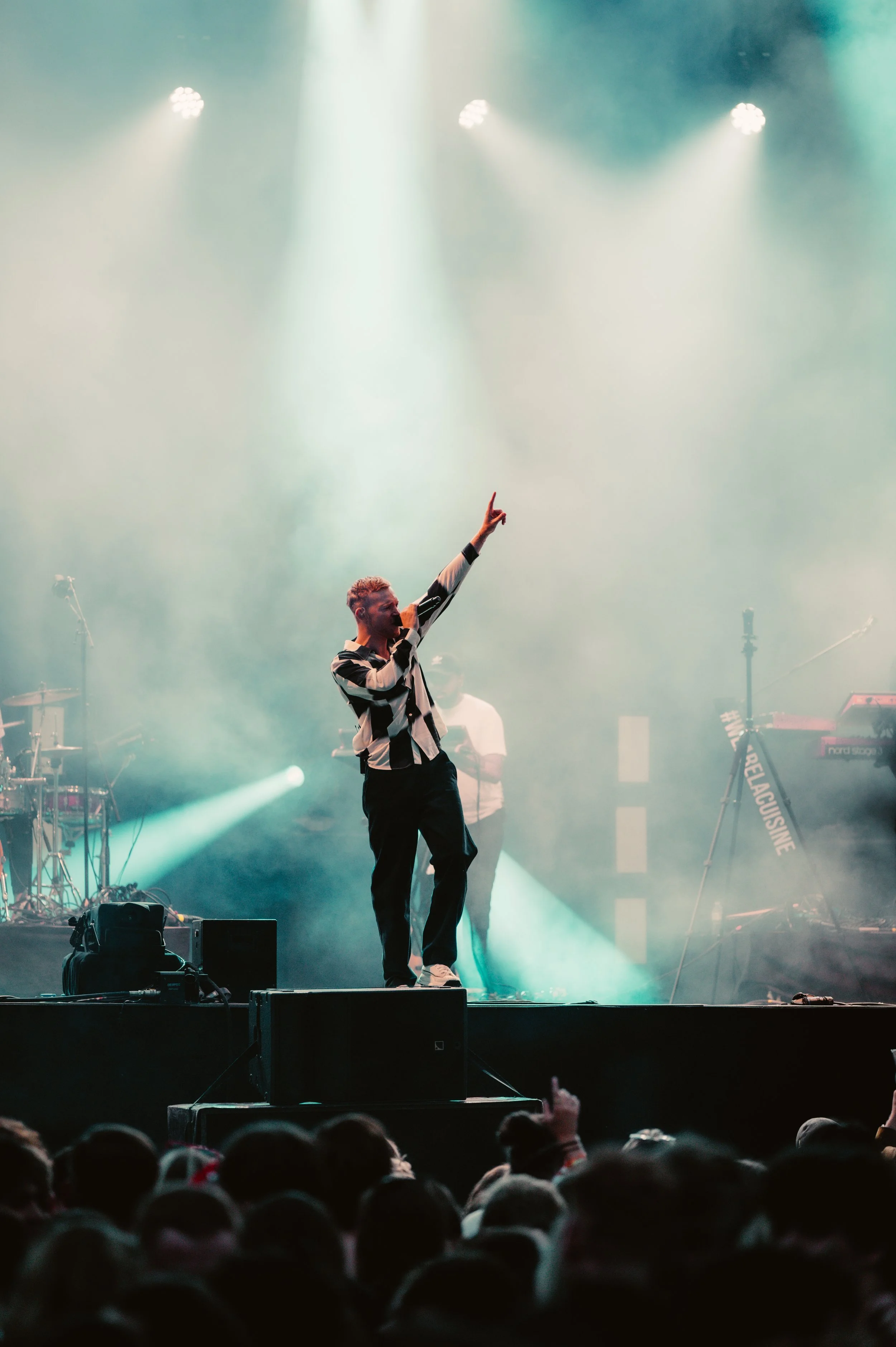 A performer on stage at a concert, holding a microphone and pointing upwards with one hand. Stage lights and smoke create a dramatic backdrop with musical equipment visible in the background.