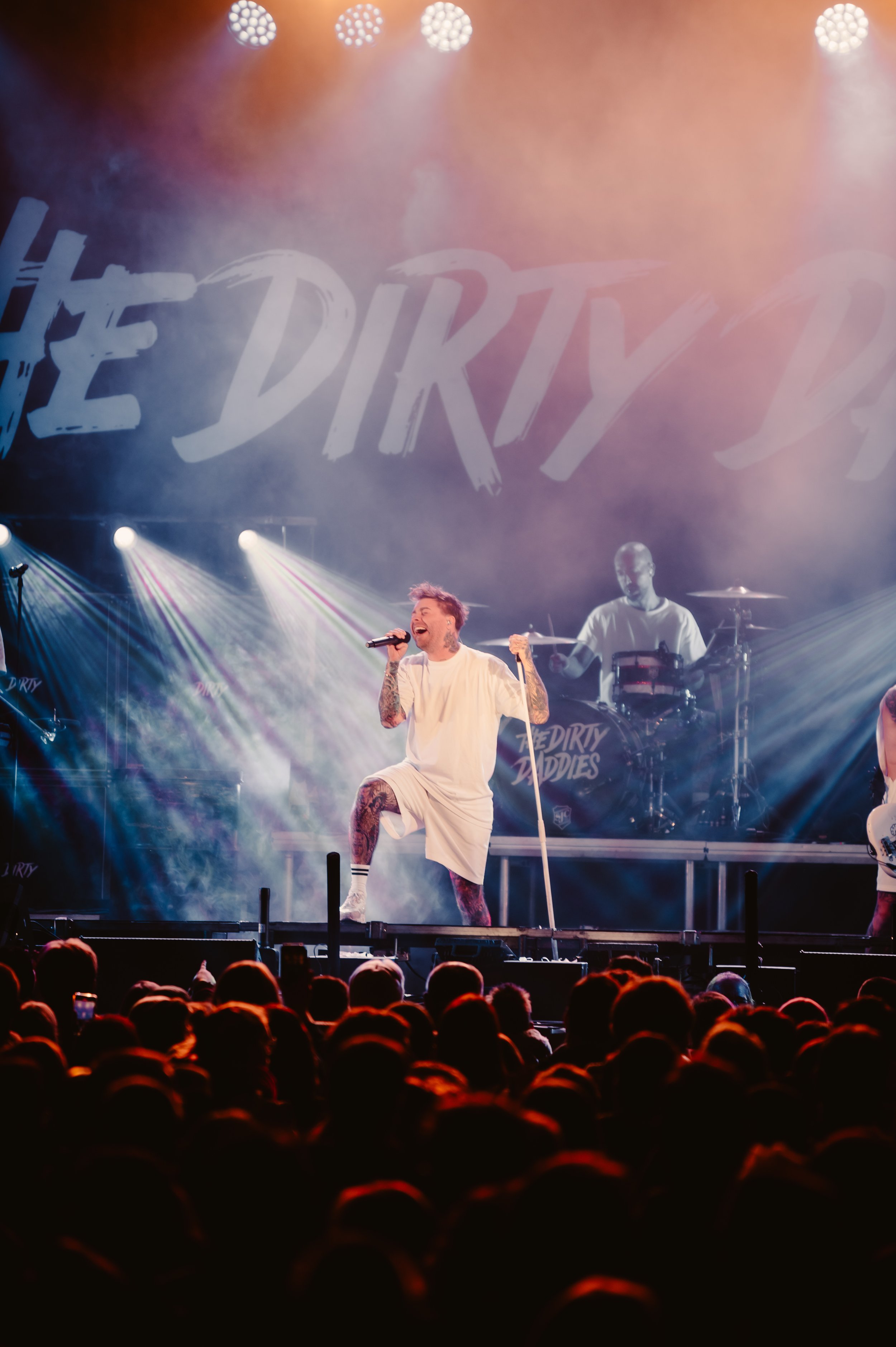 A band performing on stage with colorful lights, featuring a vocalist singing passionately into a microphone. The background displays the text 'The Dirty Daddies'. A drummer is also visible.