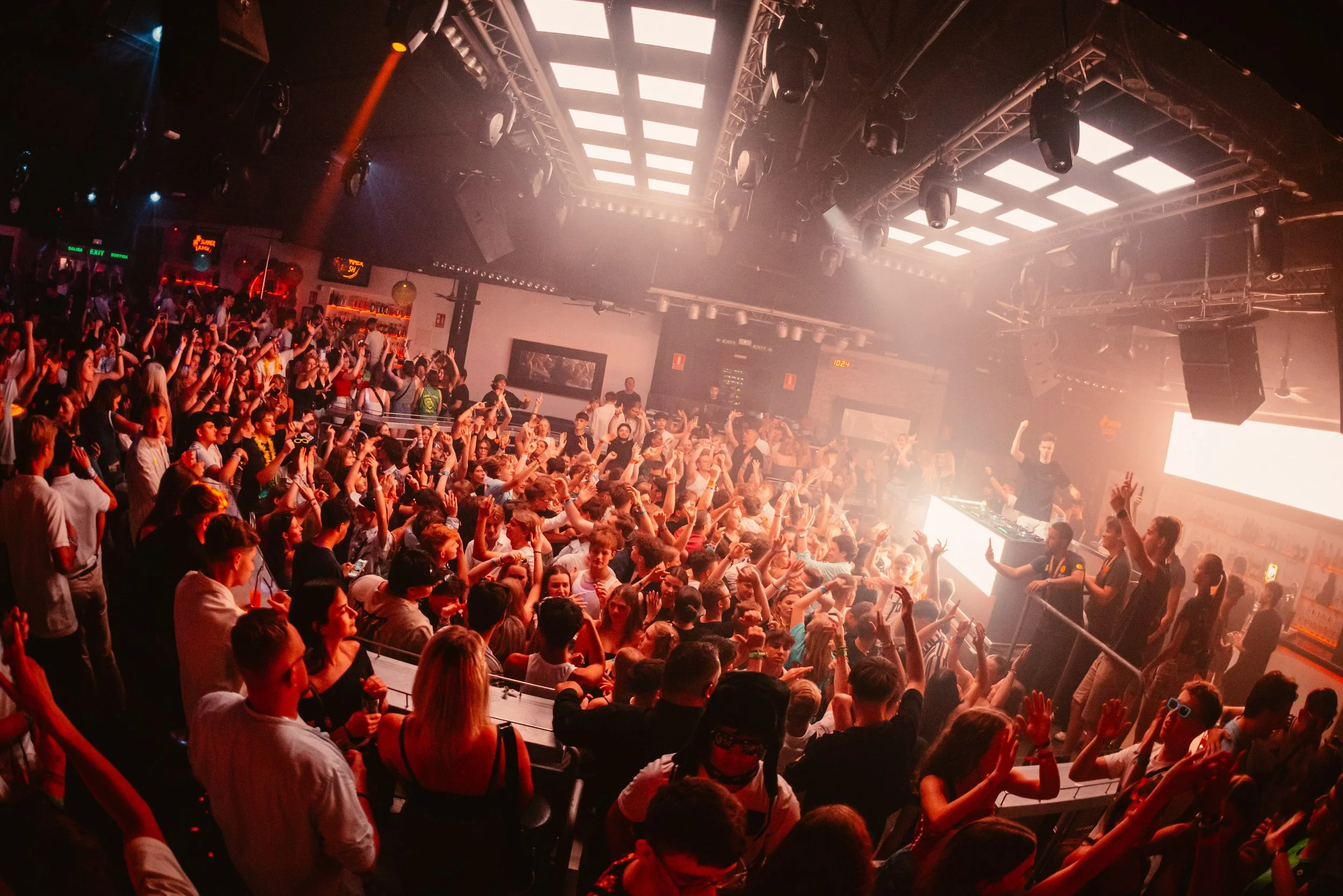 Crowded nightclub scene with people dancing and a DJ performing on stage under bright lights.
