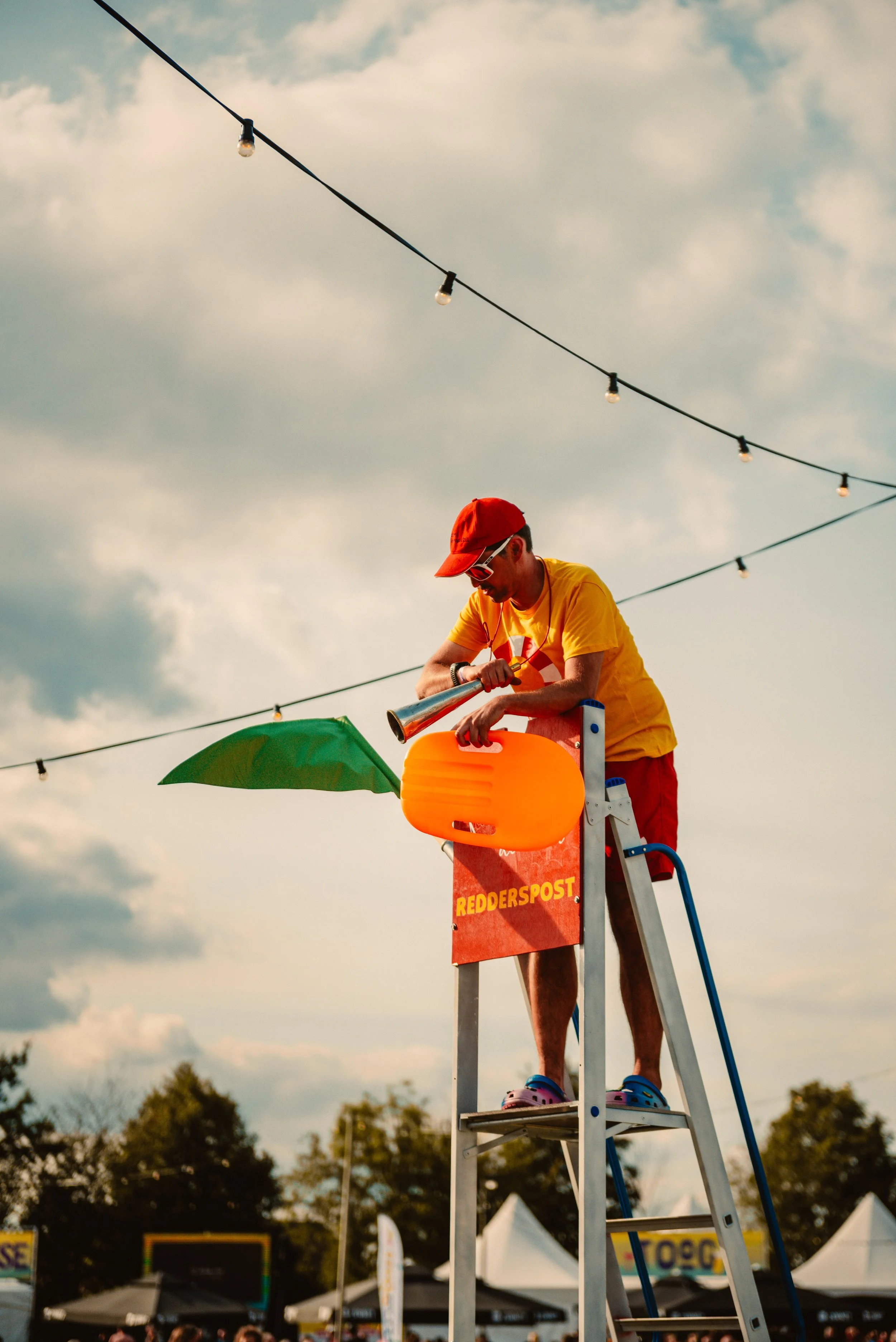 Person in colorful outfit standing on a lifeguard chair, holding a megaphone and inflatable paddle, with a green flag, outdoor event setting.