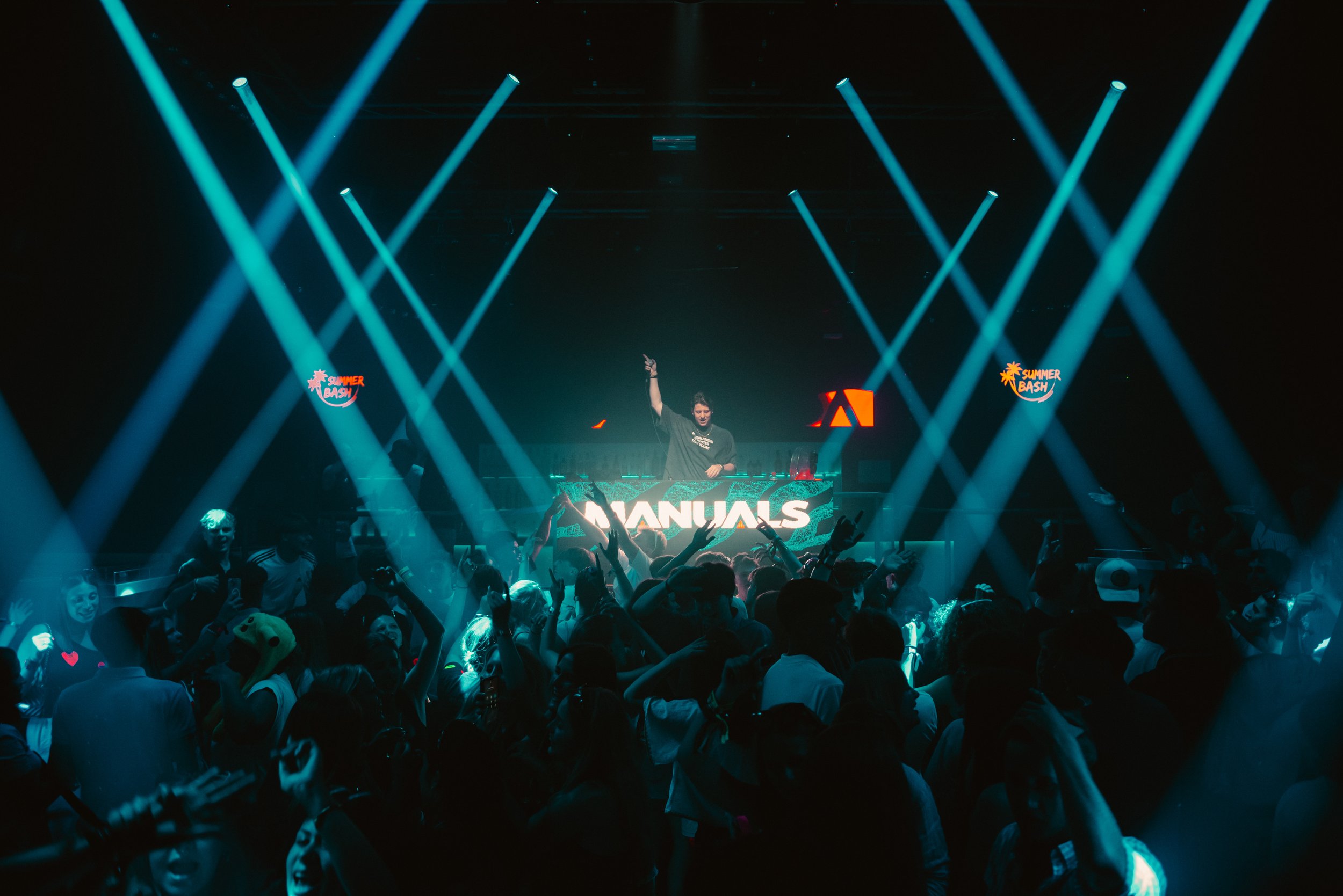 DJ performing at a nightclub with a crowd dancing, illuminated by blue and white spotlights, with screens showing graphics and signs like 'Summer Bash' and 'Manuals.'