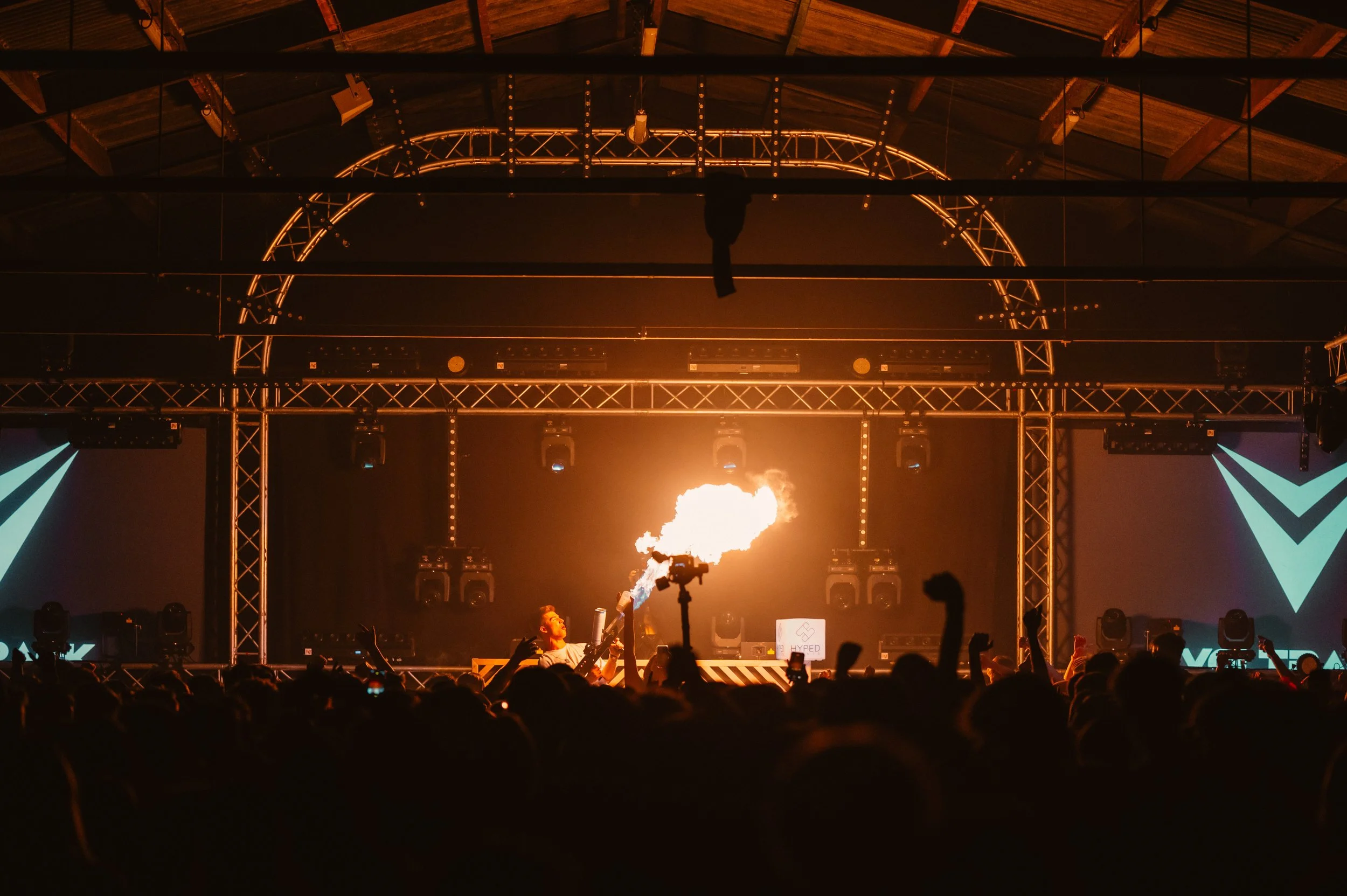 Concert scene with performer using a flamethrower on a stage, surrounded by a cheering crowd with lighting and sound equipment.