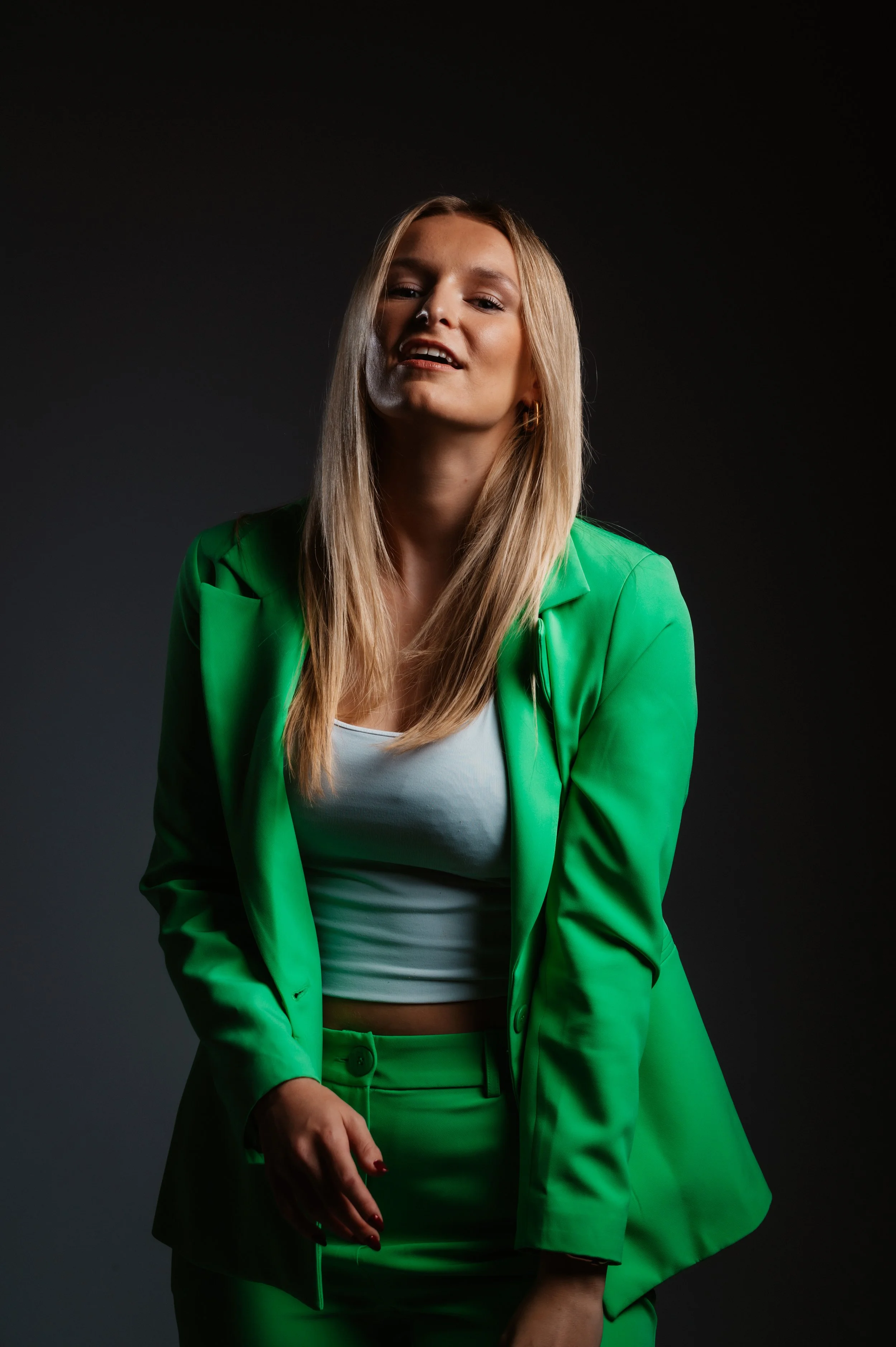 Woman wearing a bright green suit and white top against a dark backdrop, posing with a relaxed expression.