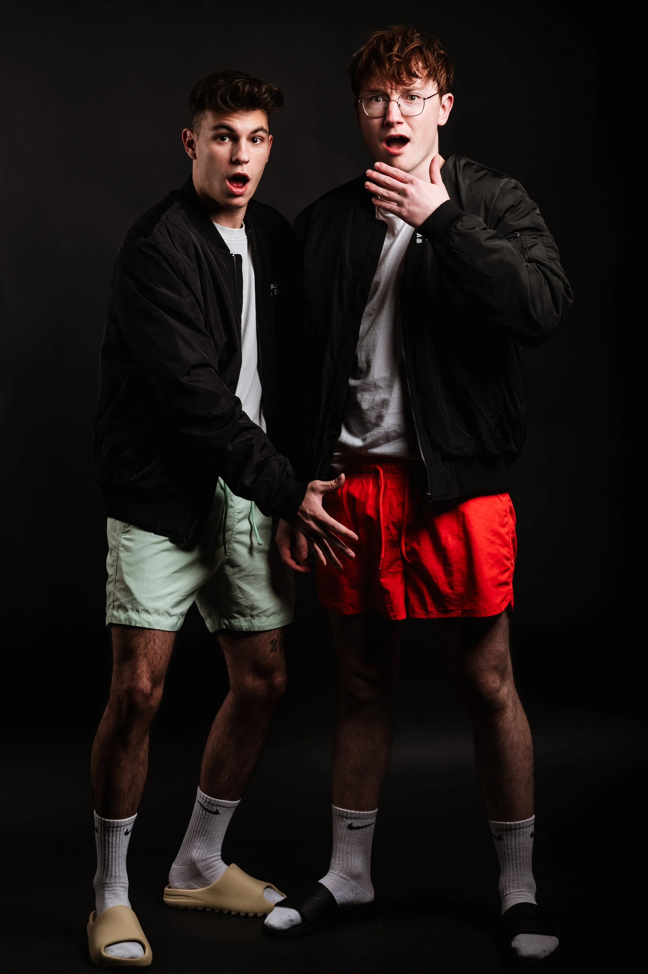 Two young men with surprised expressions, wearing black jackets, white shirts, shorts, and slides with socks, standing against a dark background.