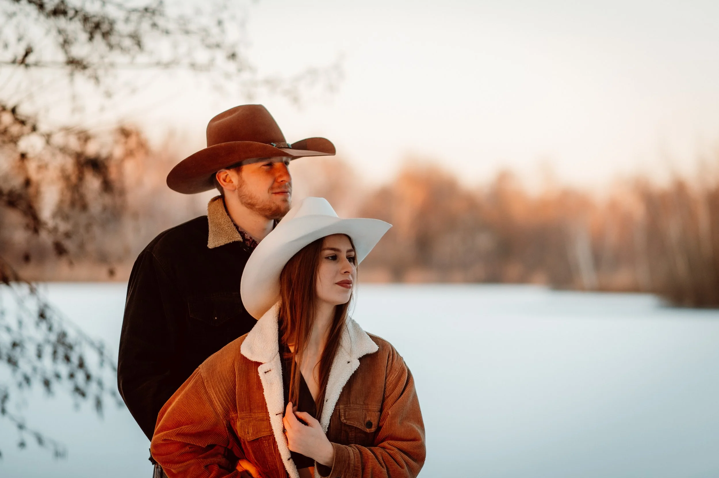 A couple wearing cowboy hats and winter coats standing outdoors near a frozen lake with trees in the background during sunset.