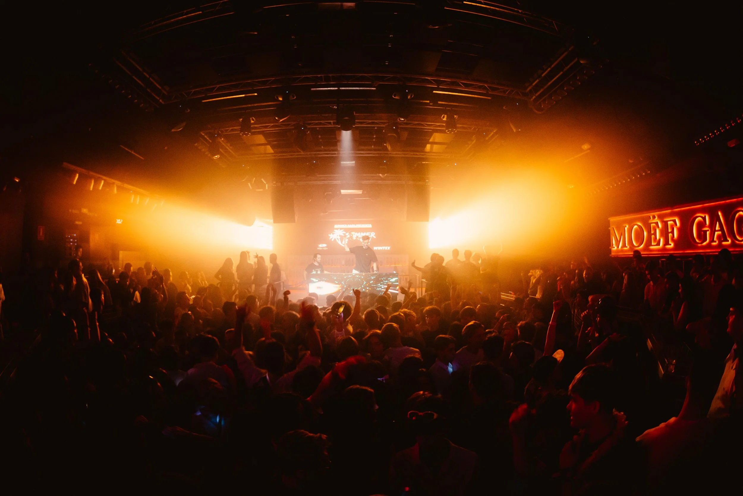 Crowded nightclub with DJ performance, bright orange lighting, and a neon "MOËF GAC" sign on the wall.