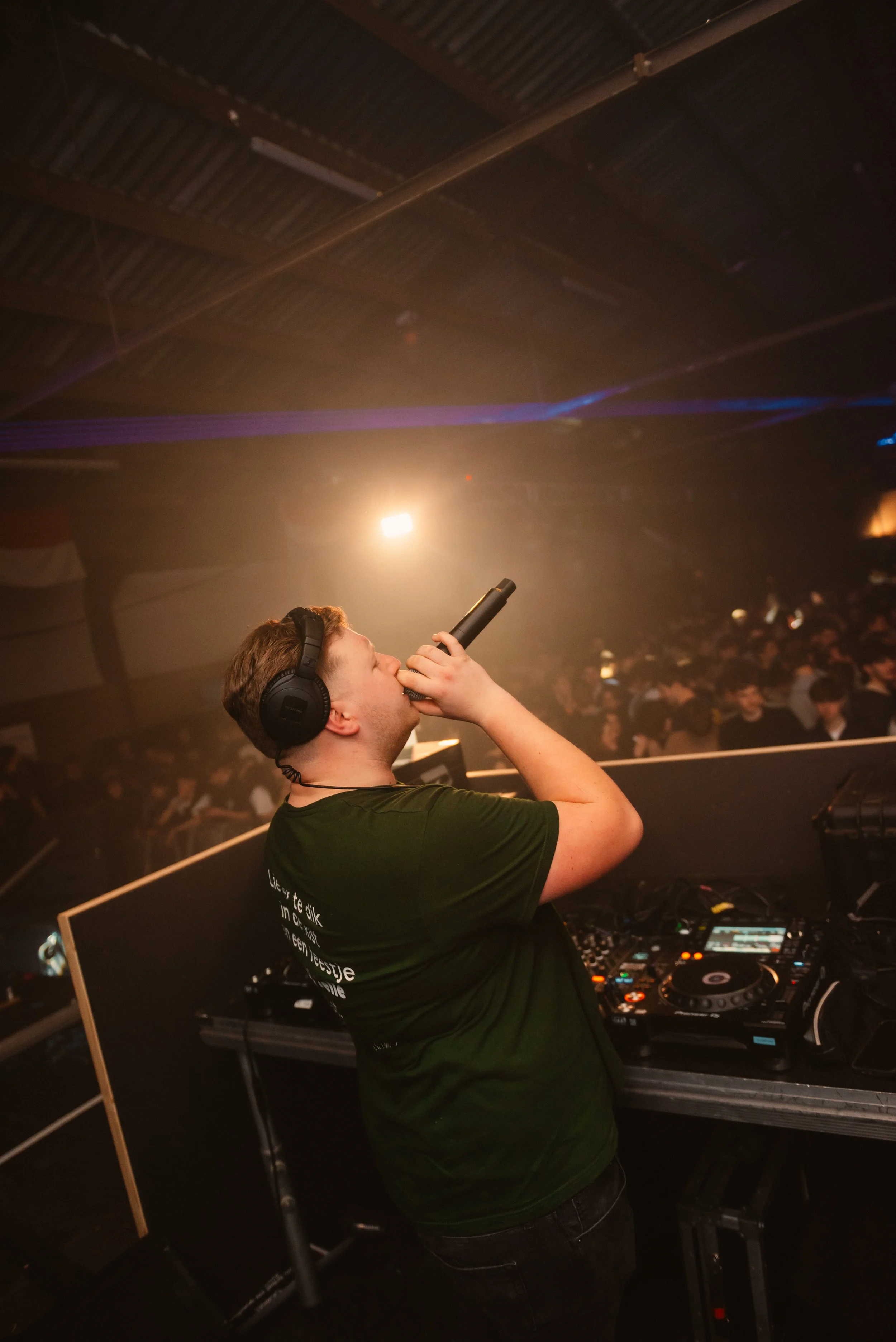DJ performing in a nightclub with a microphone, wearing headphones, standing at a DJ booth with turntables and equipment, facing a large crowd of people on the dance floor.
