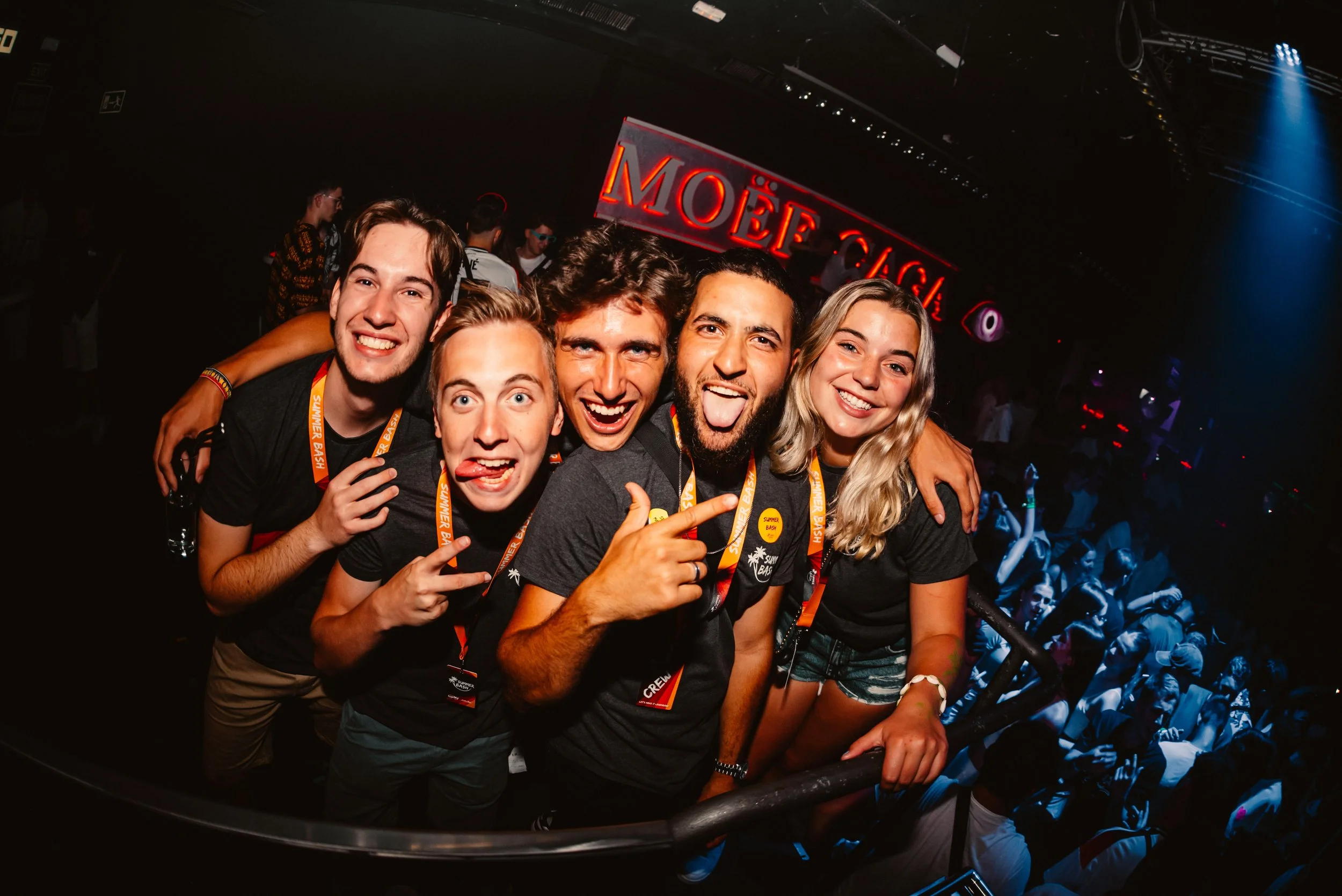 Group of five friends smiling and posing for a photo at a lively indoor party or event, wearing 'Summer Bash' lanyards. Crowd in the background suggests a festive atmosphere.