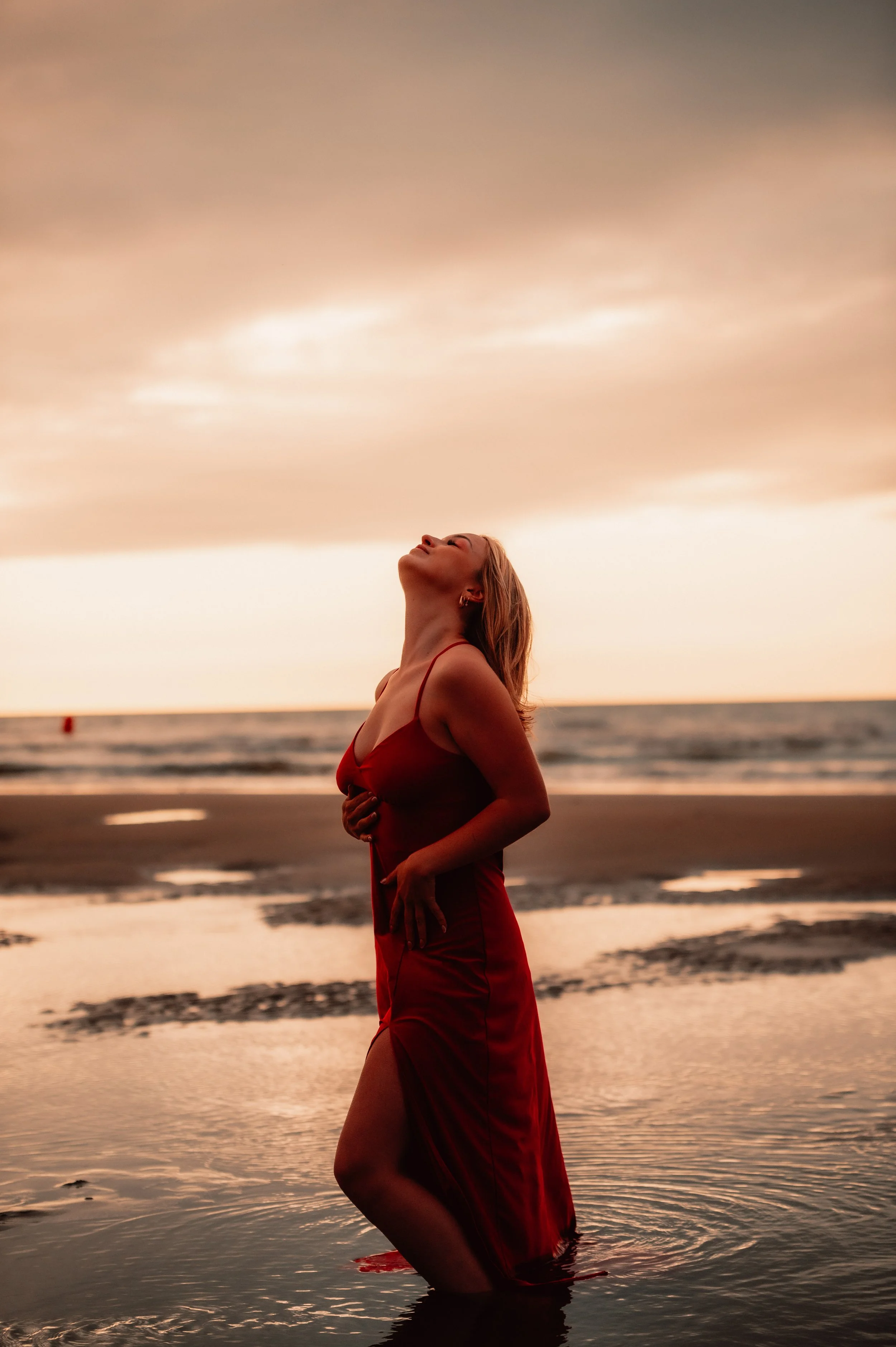 Woman in red dress standing in shallow water on beach at sunset
