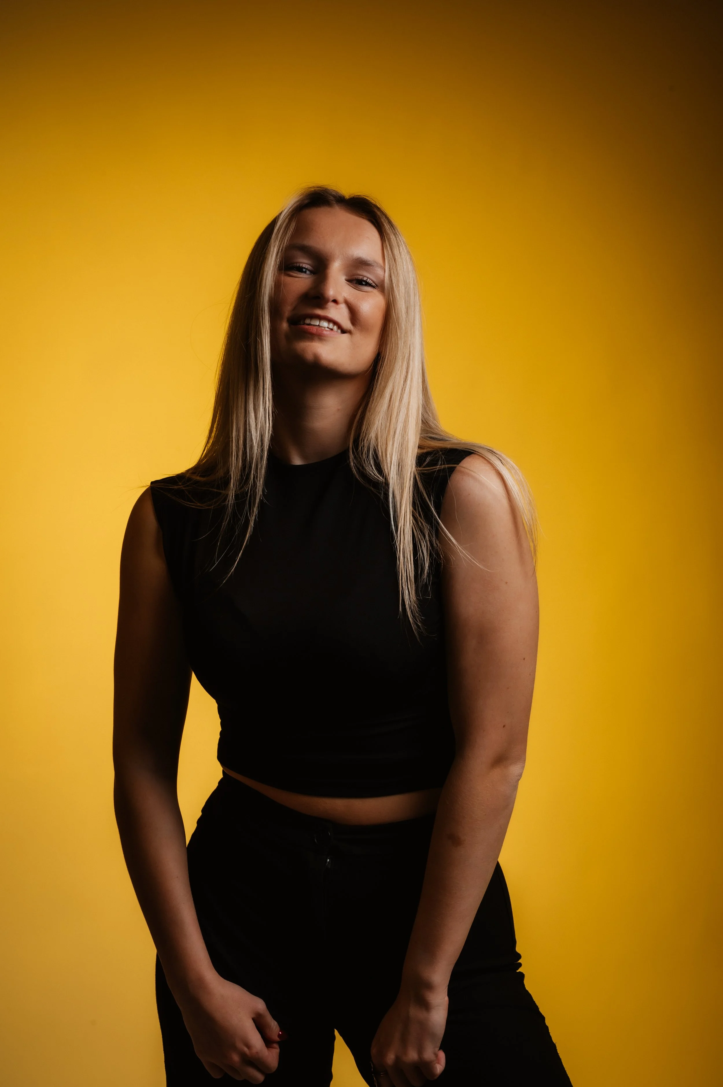 Smiling woman in black outfit with long blonde hair on yellow background.