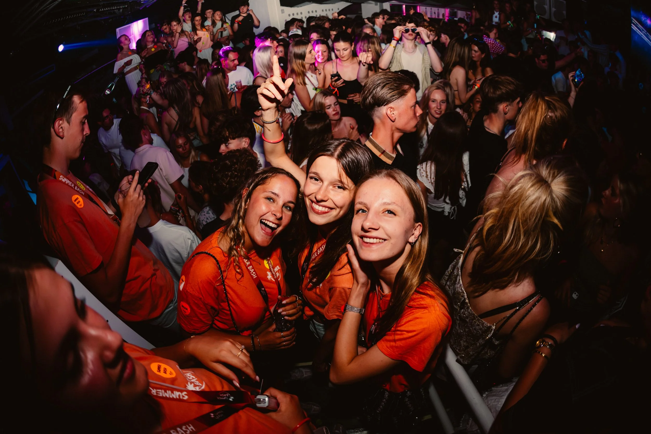 Crowd at a lively party with people smiling and taking selfies, wearing orange shirts, under colorful lights.