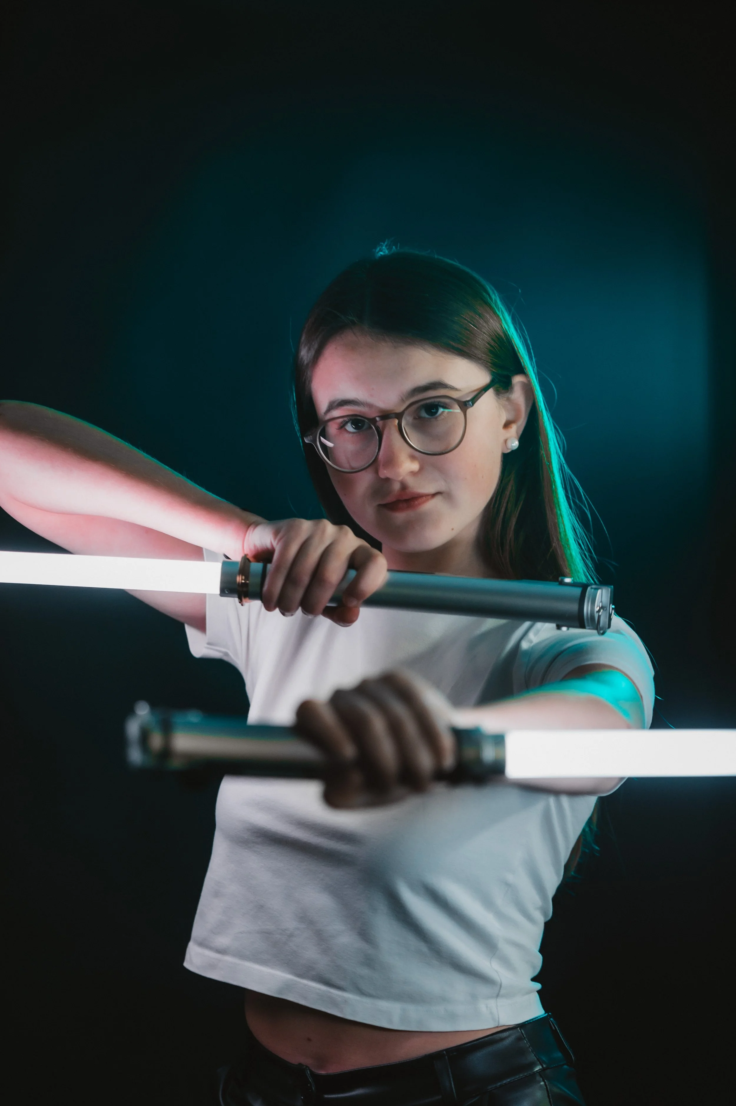 Person wearing glasses and holding two light sticks in a dimly lit environment, dressed in a white shirt.