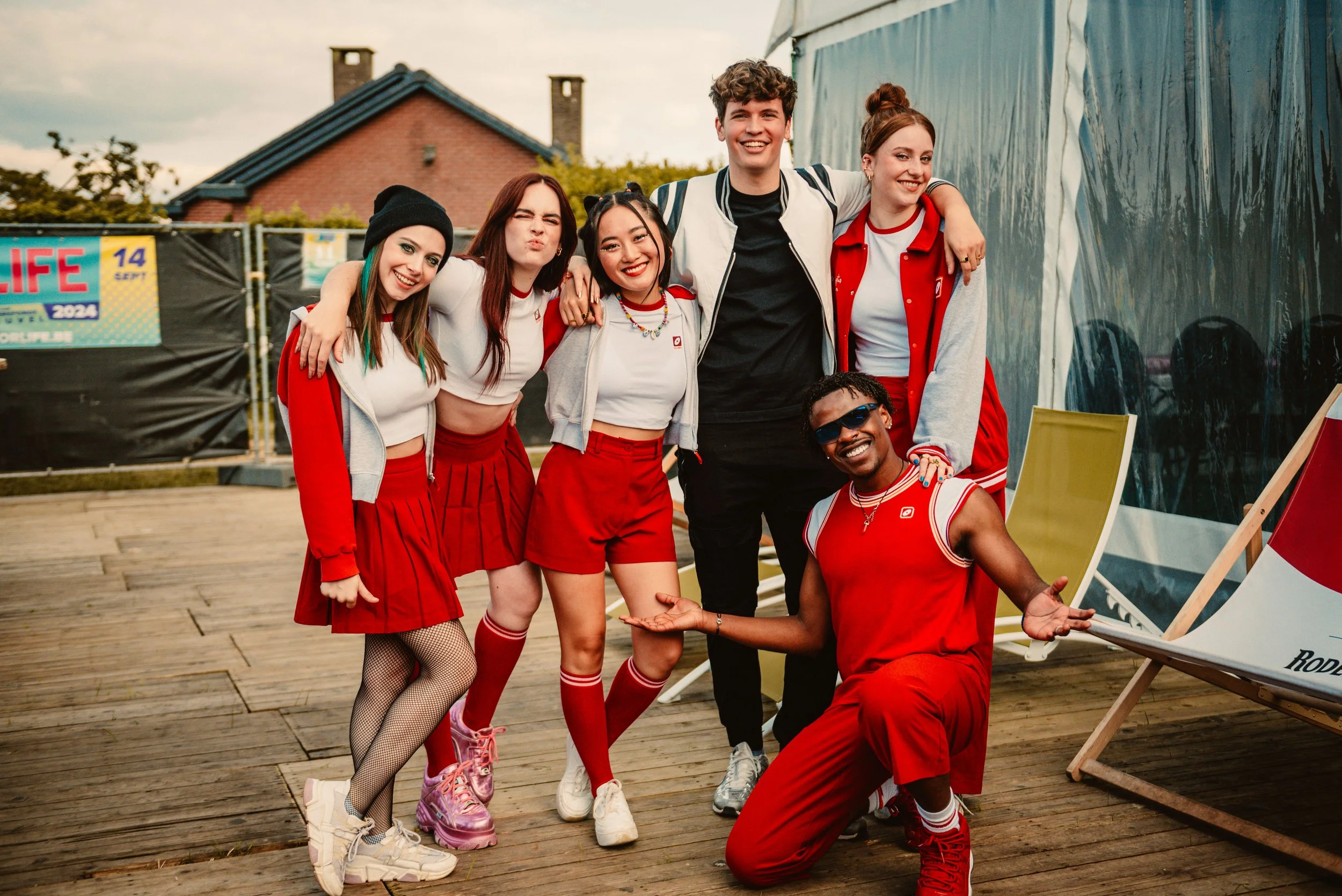 Group of young adults in matching red and white outfits posing outdoors, smiling and making playful gestures, with a building and event banners in the background.
