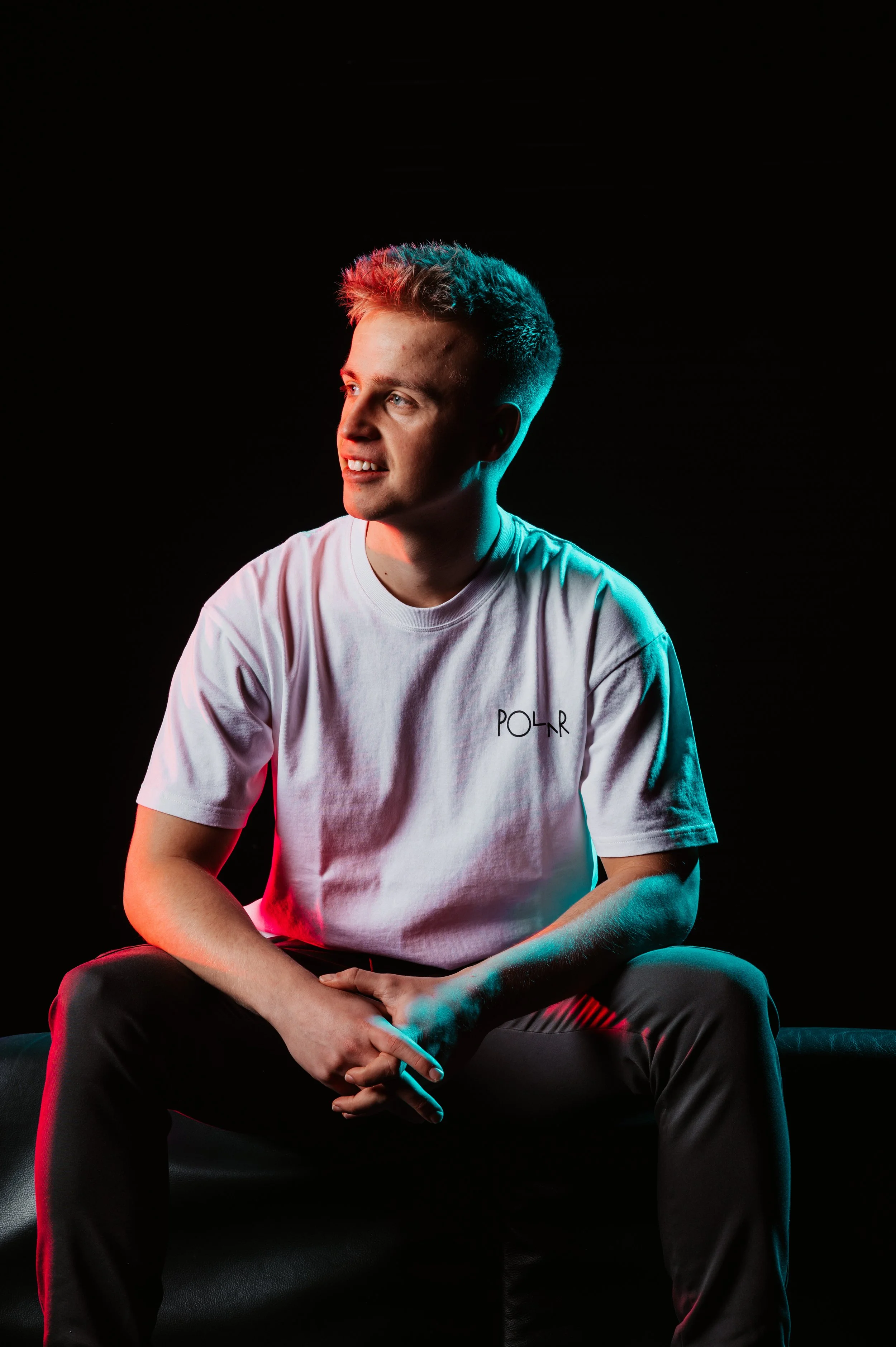 Person sitting on a black surface, wearing a white t-shirt with "POLR" text, lit by colored lights in a dark setting.
