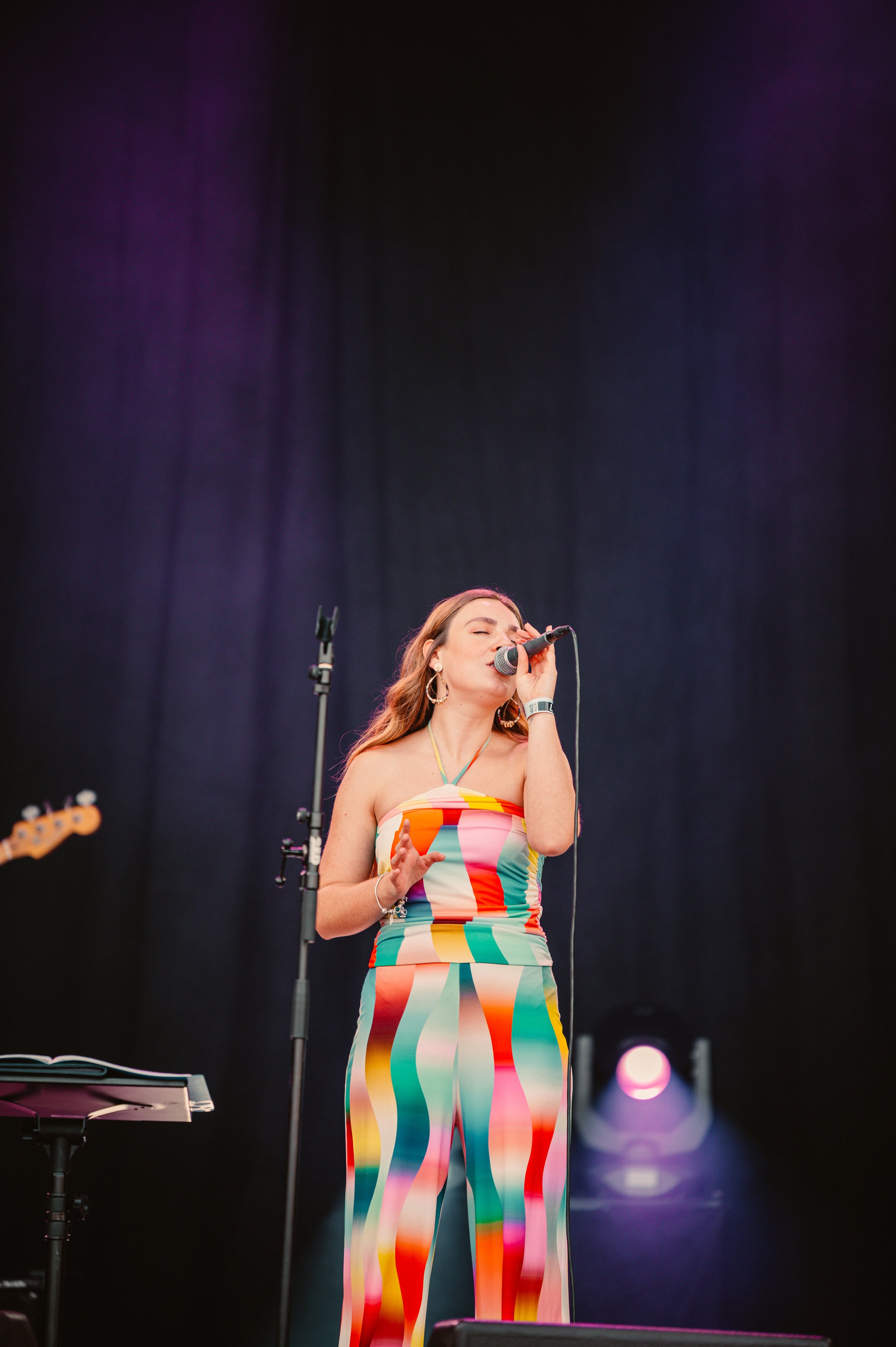 Female singer performing on stage, wearing a colorful striped outfit, holding a microphone with closed eyes and a music stand nearby.