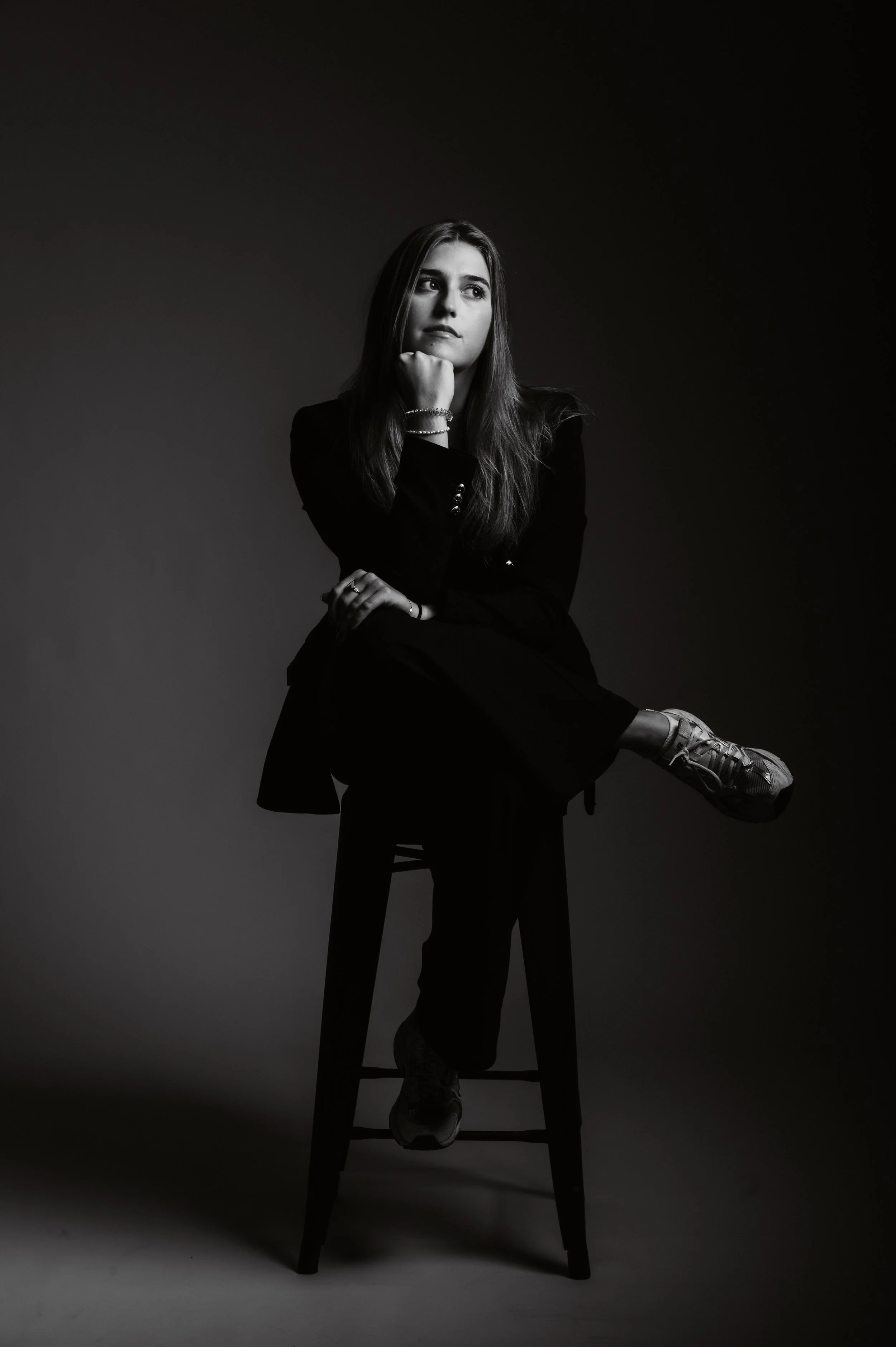 Black and white photo of a woman in a suit sitting on a stool, resting her chin on her hand and looking thoughtful.