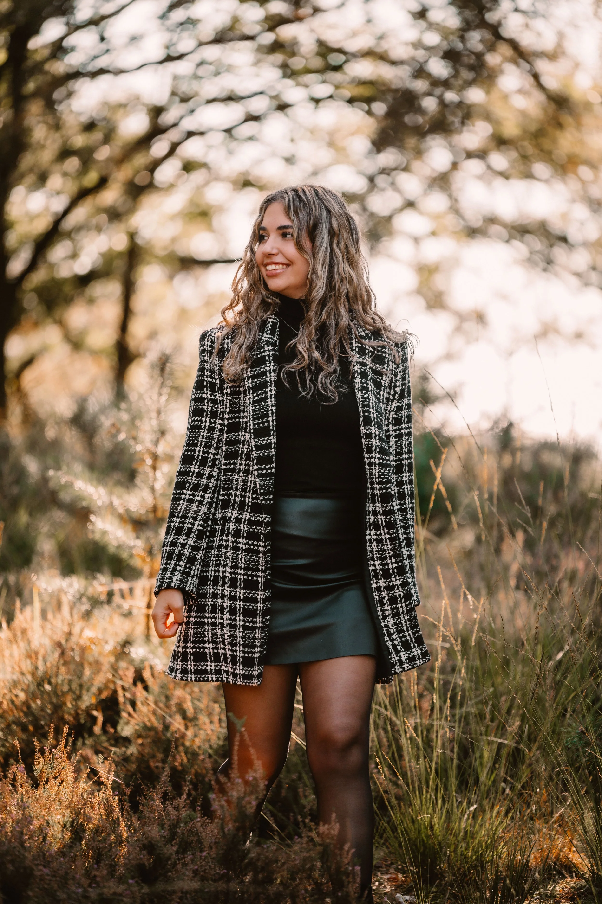 Woman in black and white checkered coat, walking in nature, smiling, during autumn.