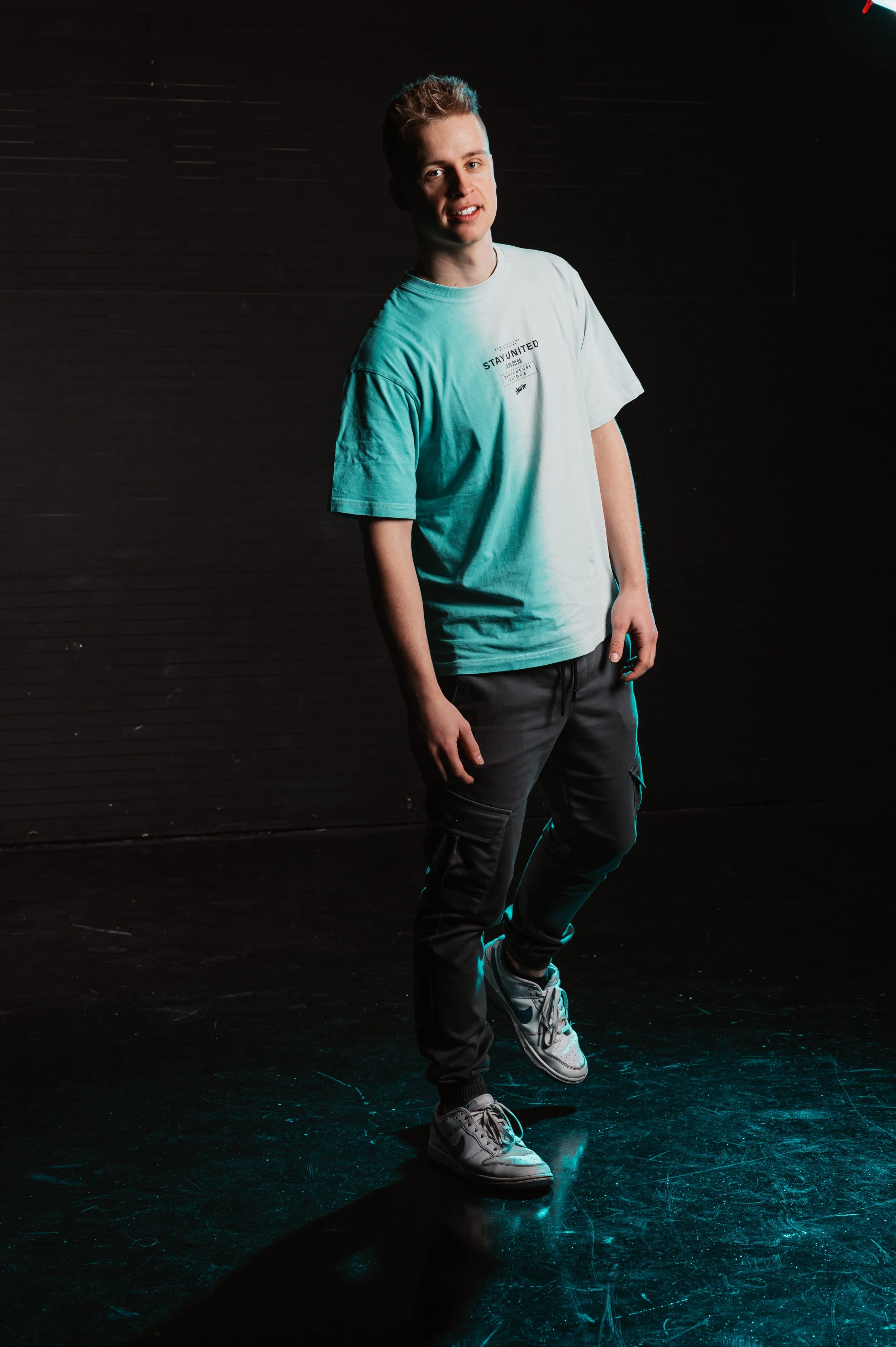 Person wearing a light blue shirt and black pants standing on a dark stage, under studio lighting.