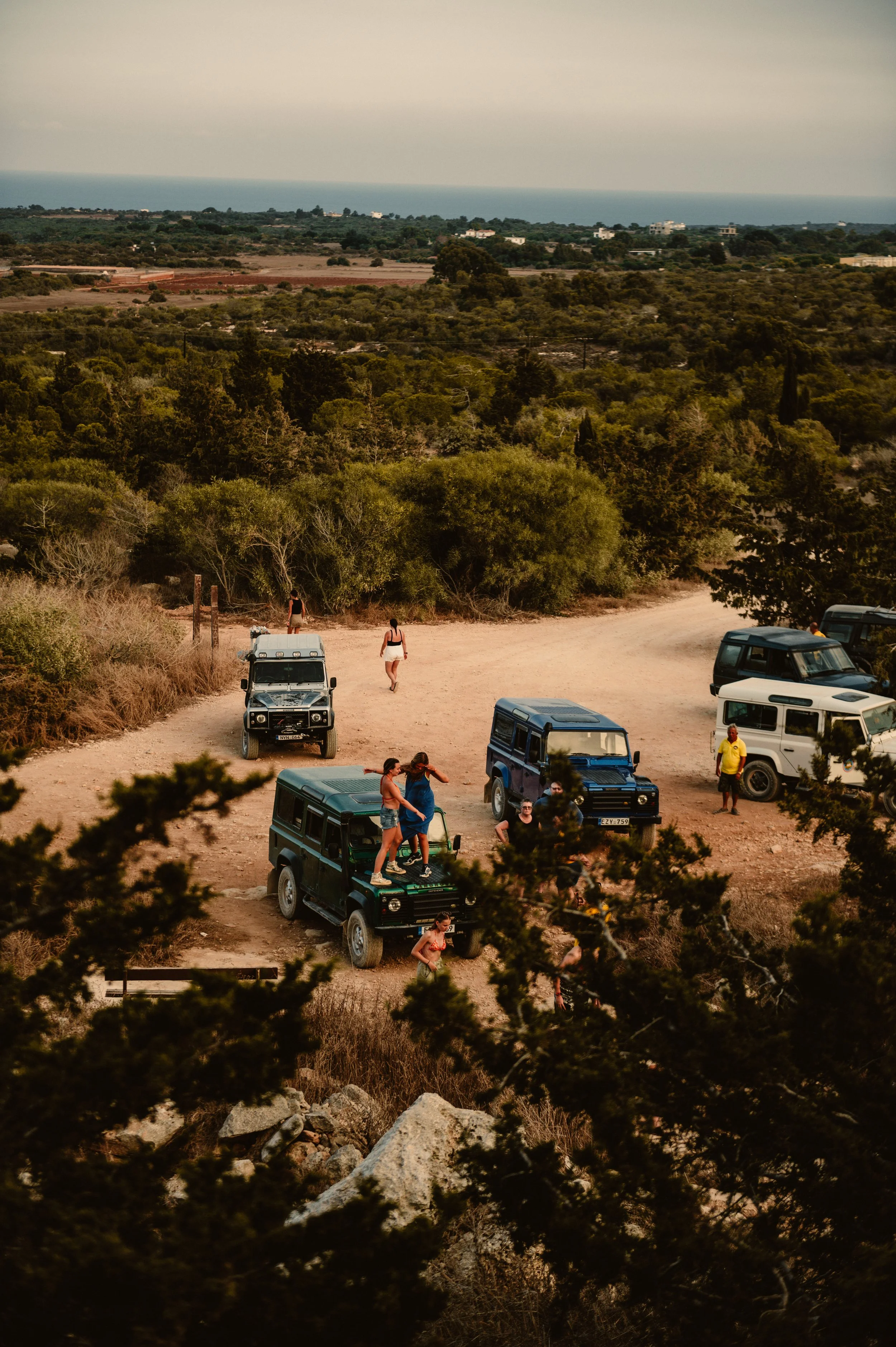Aerial view of people gathered around several off-road vehicles in a scenic, rural area with greenery and distant horizon.
