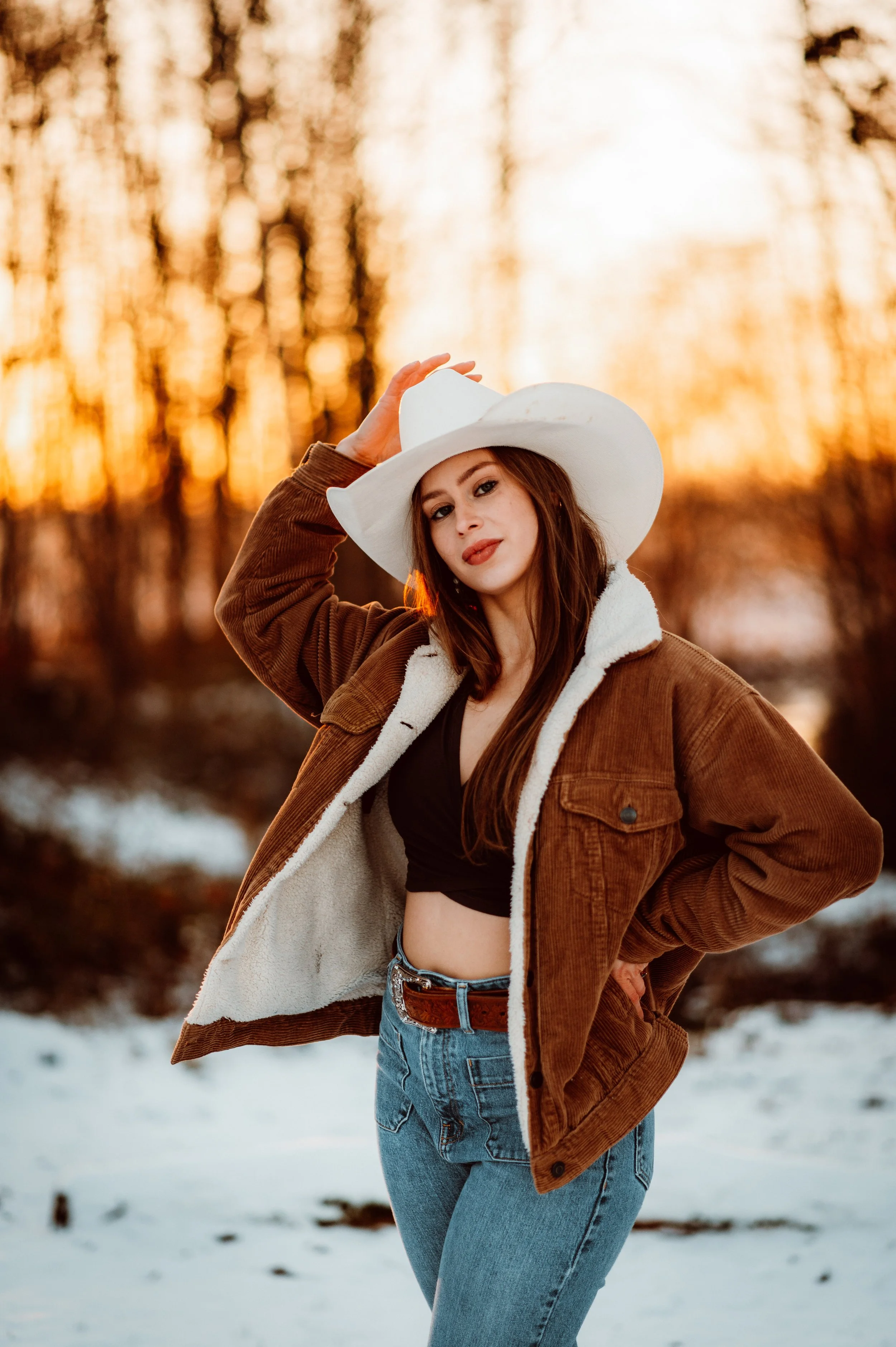 A woman in a brown jacket and a white cowboy hat poses outdoors during sunset, standing in snow with trees in the background.