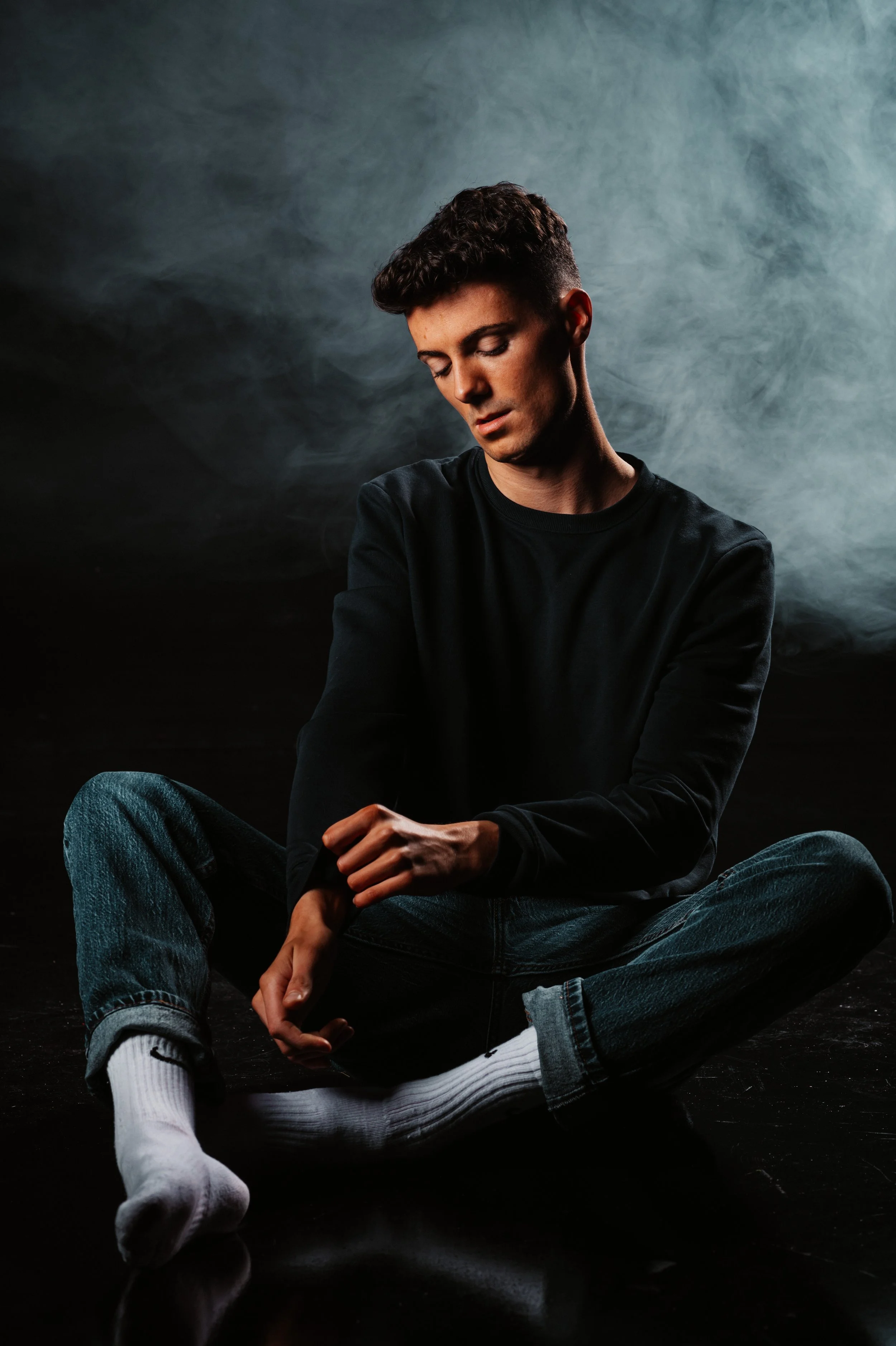 Young man in dark clothing sitting cross-legged on the floor with a smoky background.