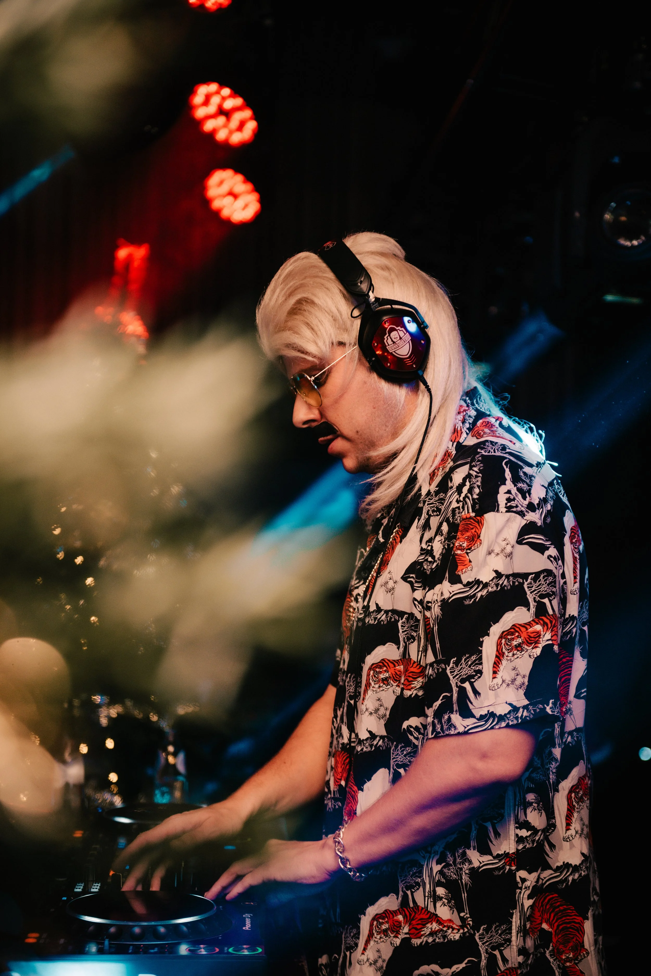DJ with blonde hair wearing headphones and a patterned shirt, mixing on DJ equipment at a club, under red stage lights.