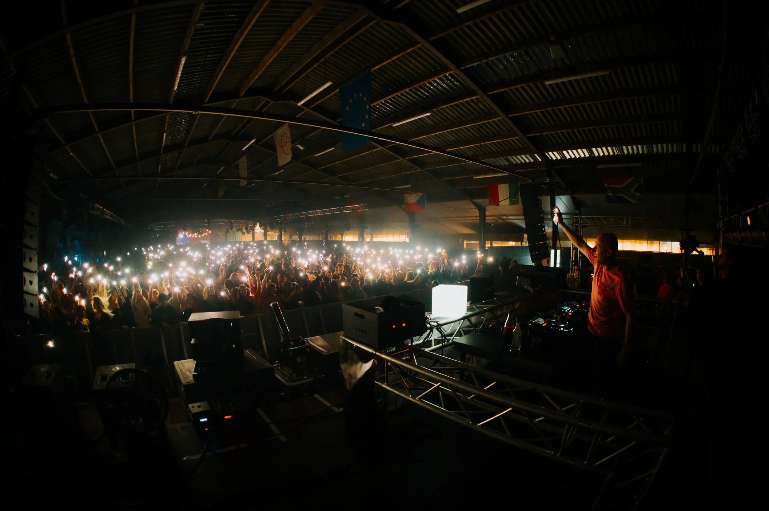 DJ performing on stage with audience holding up smartphone lights in a dimly lit venue.