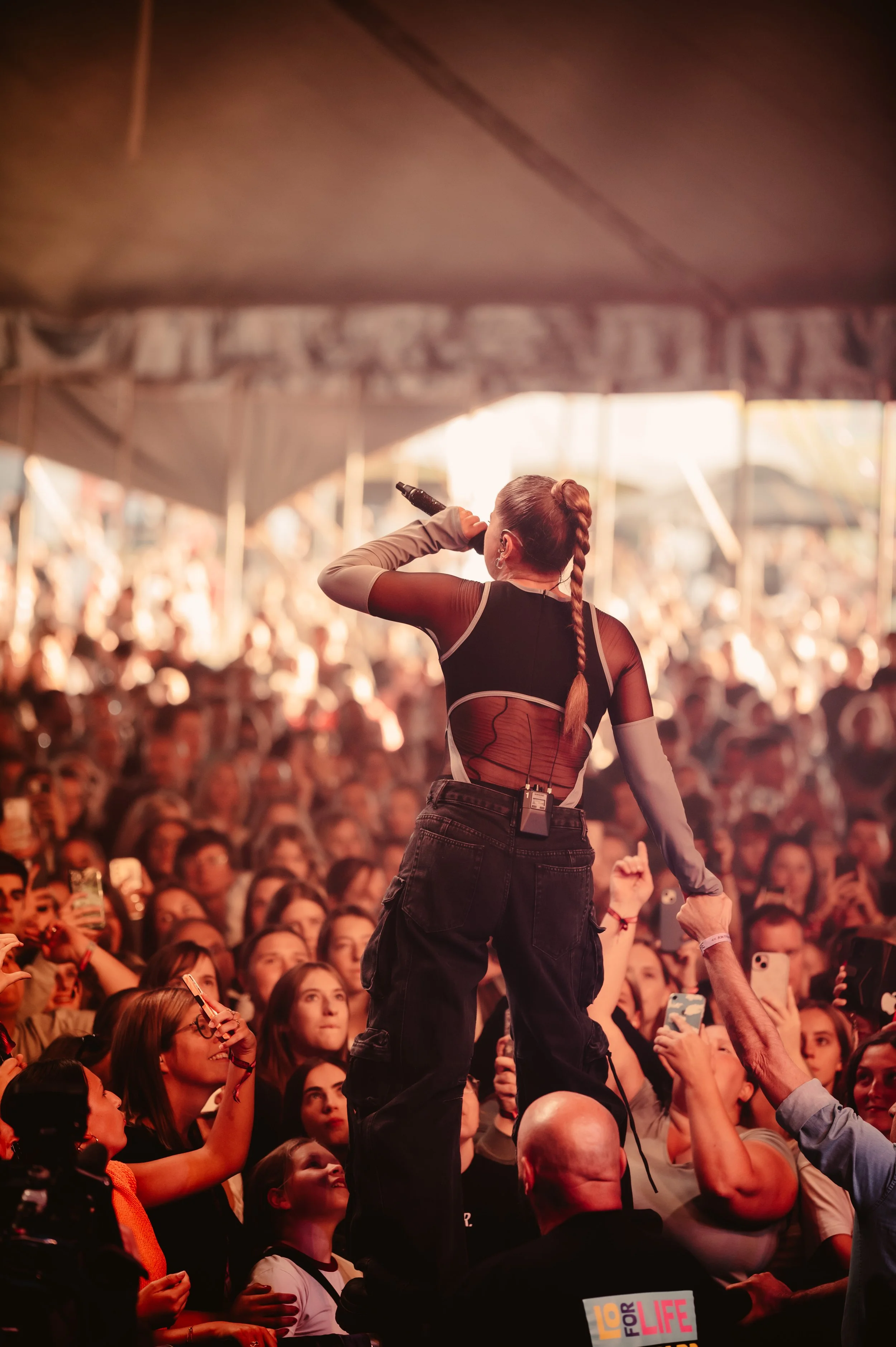 Singer performing on stage in front of a large crowd at a concert, holding a microphone and making contact with fans, under a tent.