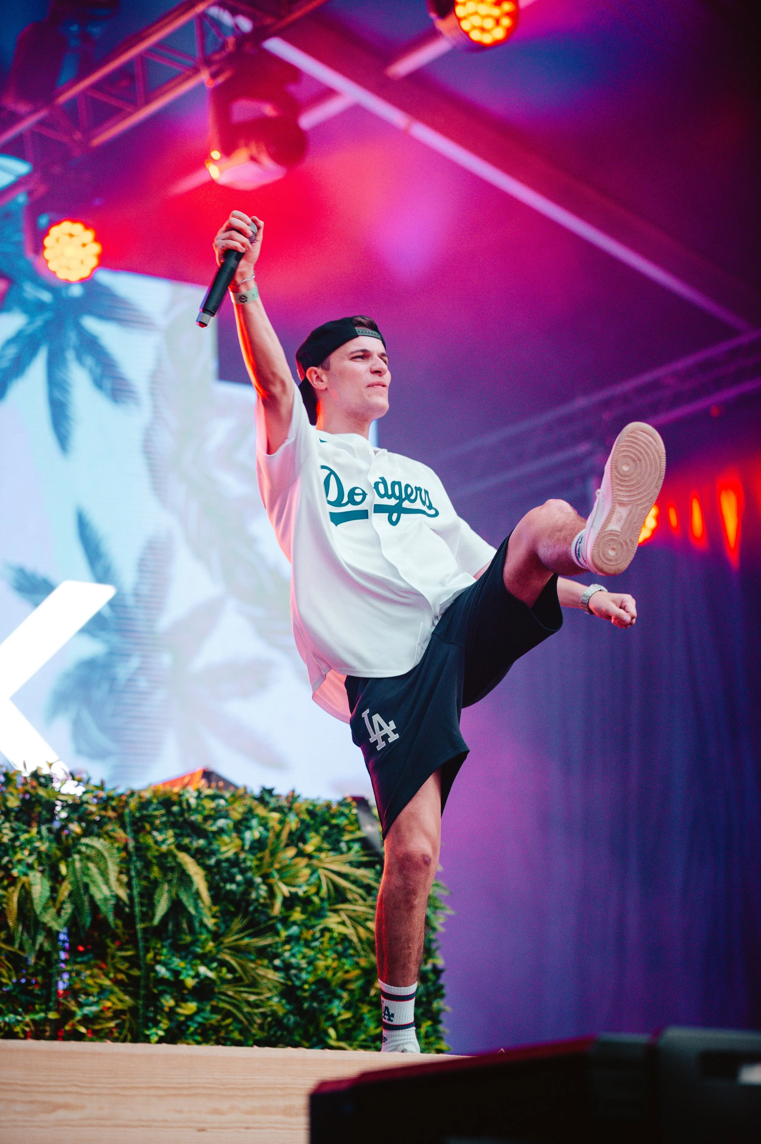 Performer on stage wearing a Dodgers shirt and cap, holding a microphone, with colorful stage lights and screen with palm leaves in the background.