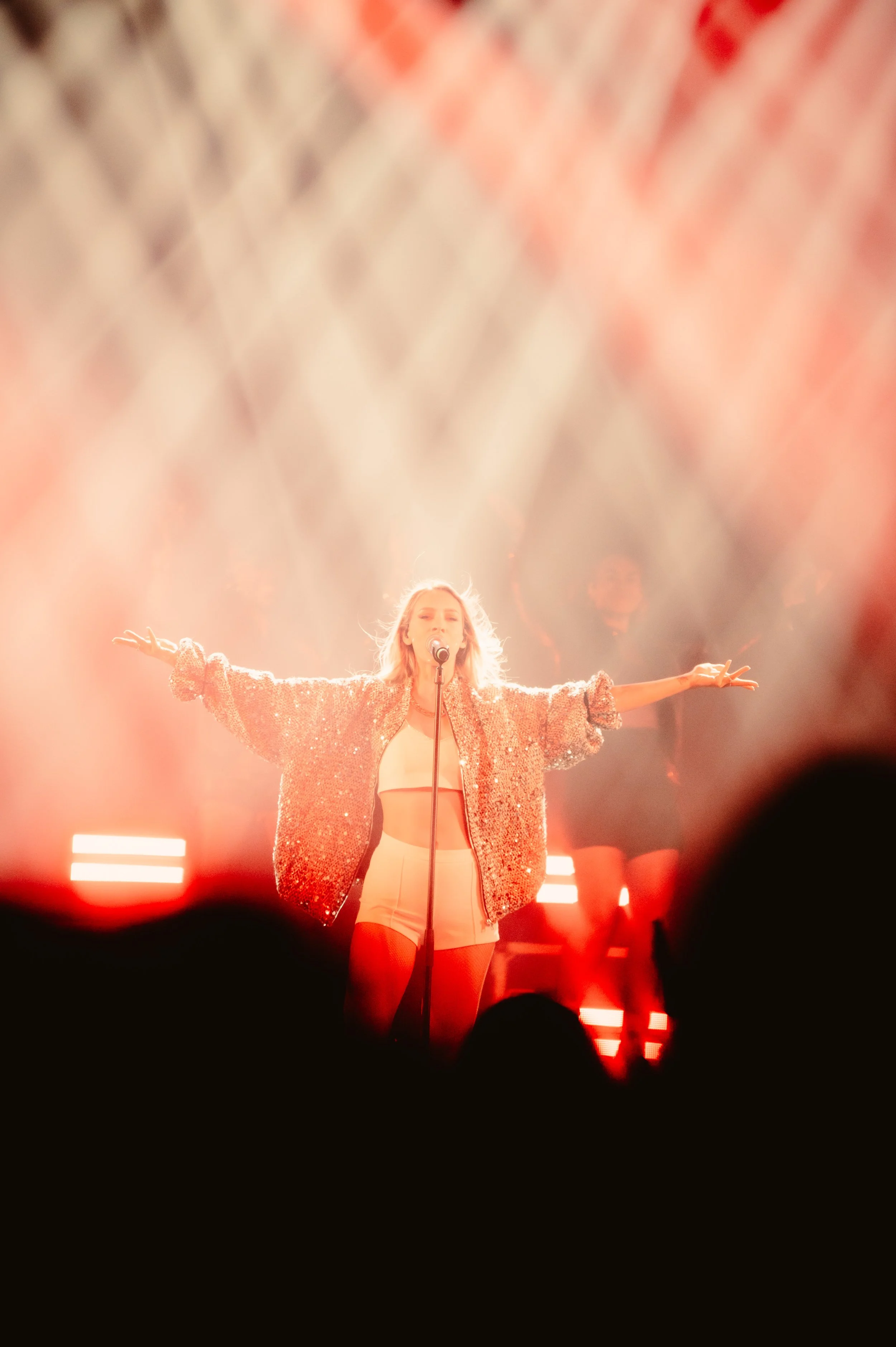 Performer singing on stage with outstretched arms, wearing a sparkling jacket and light clothing, under bright lights.