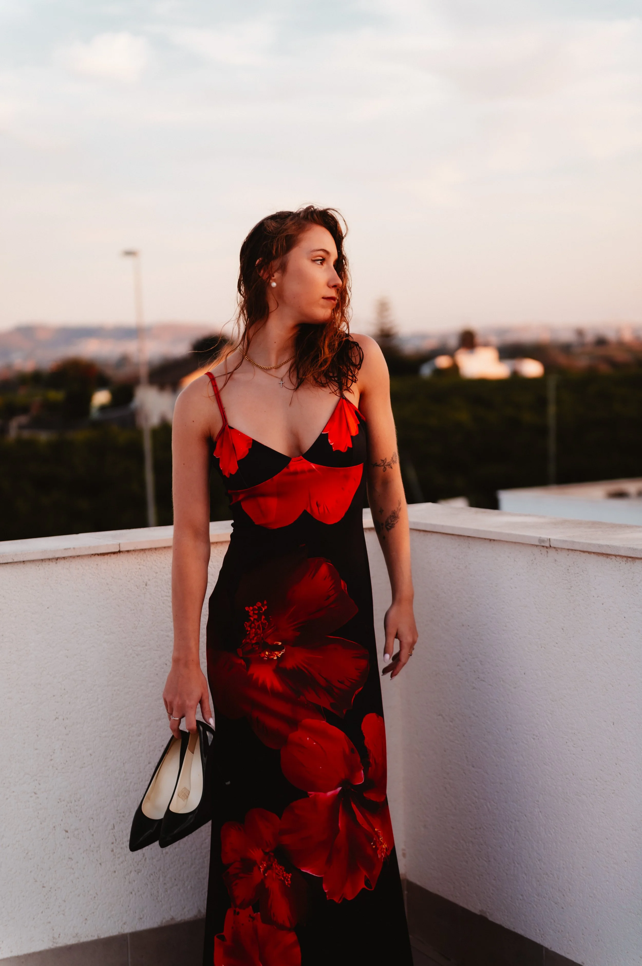 A woman in a long black evening dress with a red floral pattern, holding a pair of black and white high heels, standing on a rooftop with a cityscape in the background during sunset.