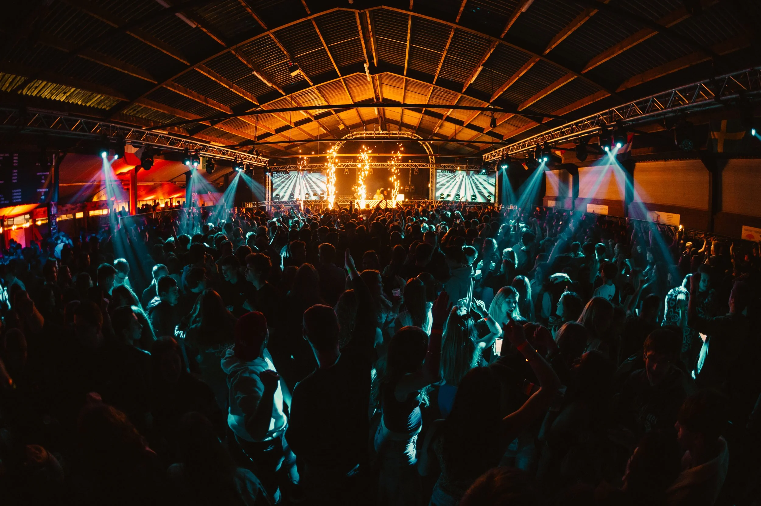 Large crowd dancing at an indoor music concert with colorful stage lights and a dynamic atmosphere.