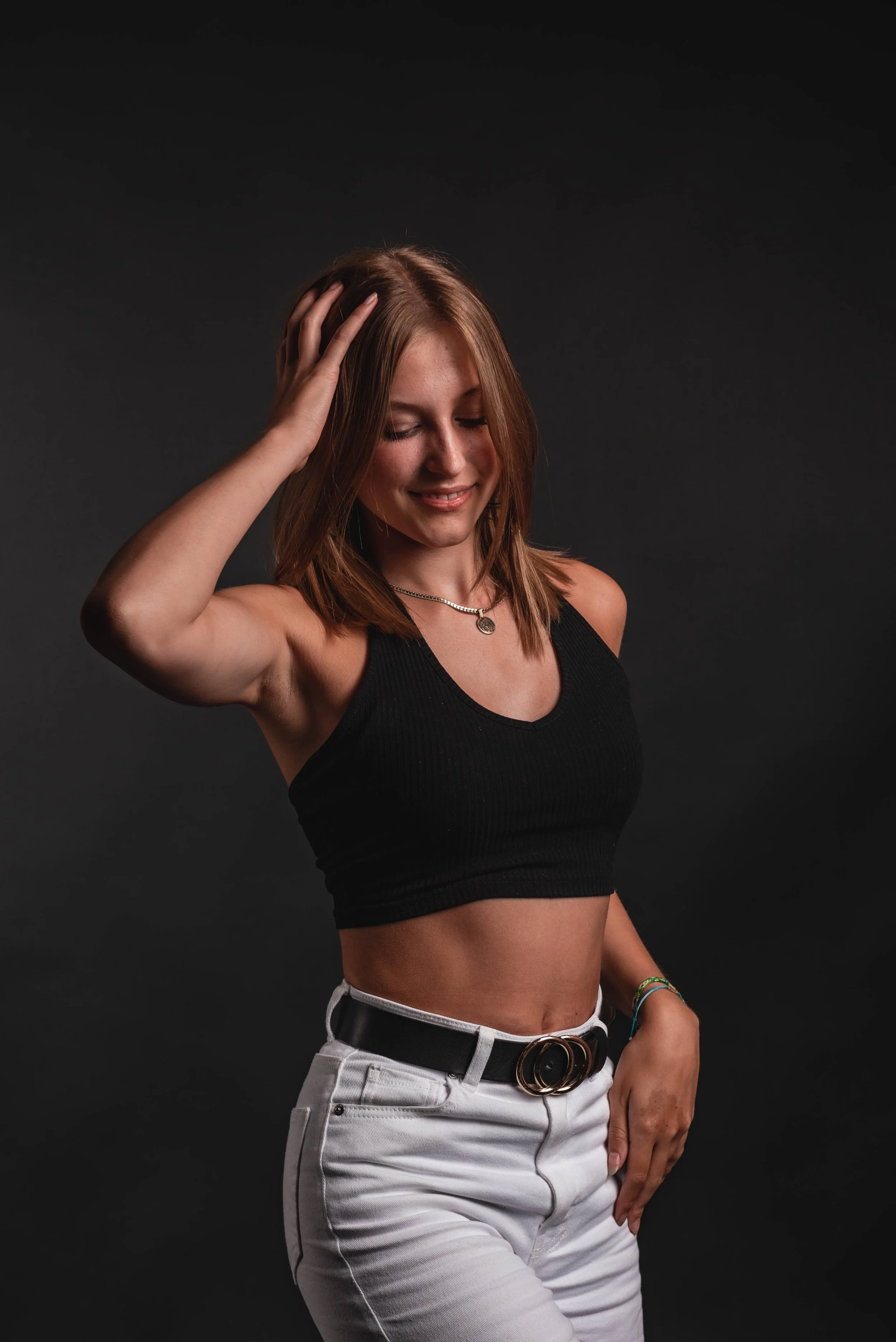 Woman in black crop top and white pants with black belt, posing with hand on head, against a dark background.