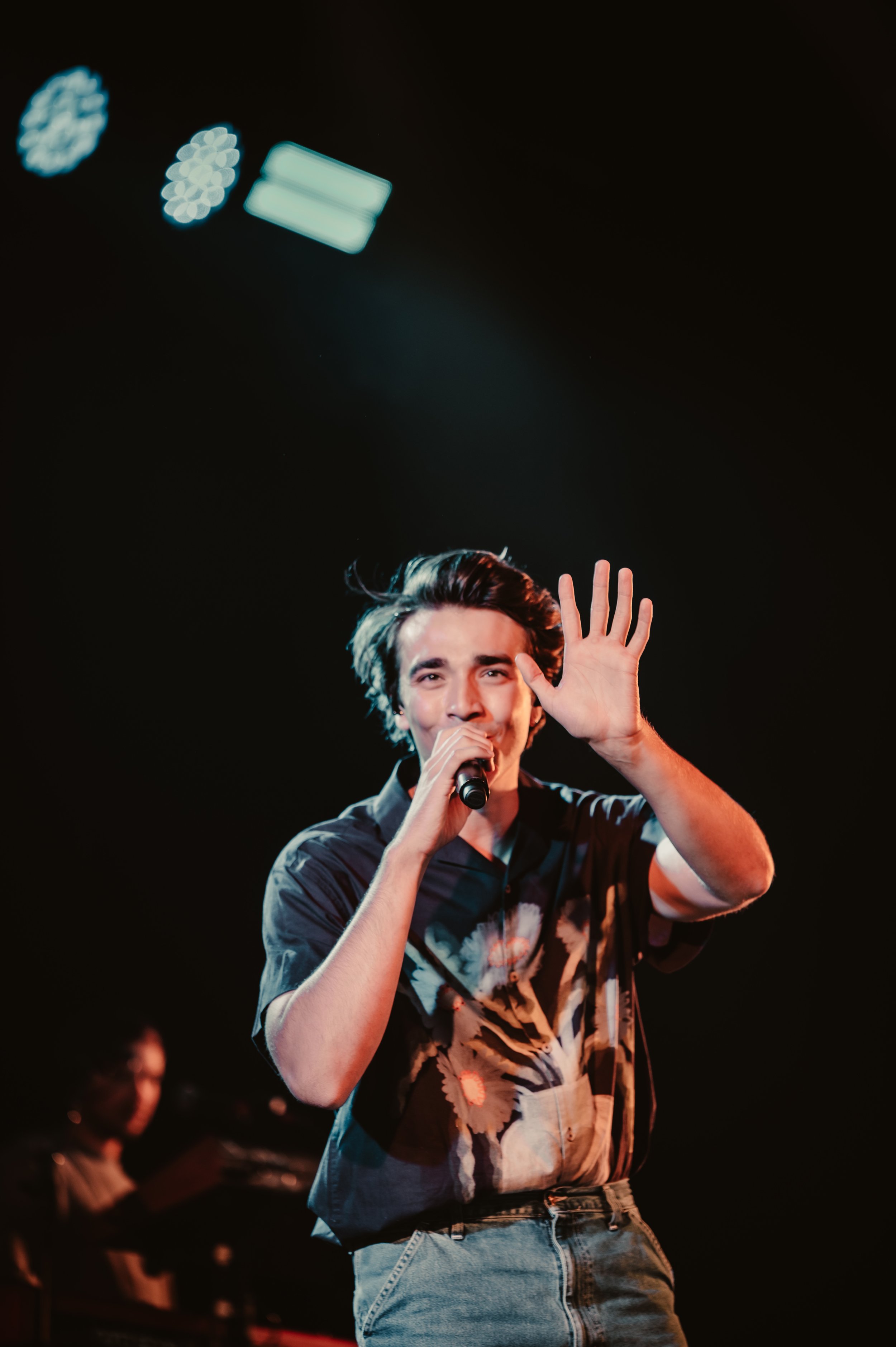 Young male singer performing on stage with microphone, wearing a patterned shirt and jeans, with stage lights in the background.