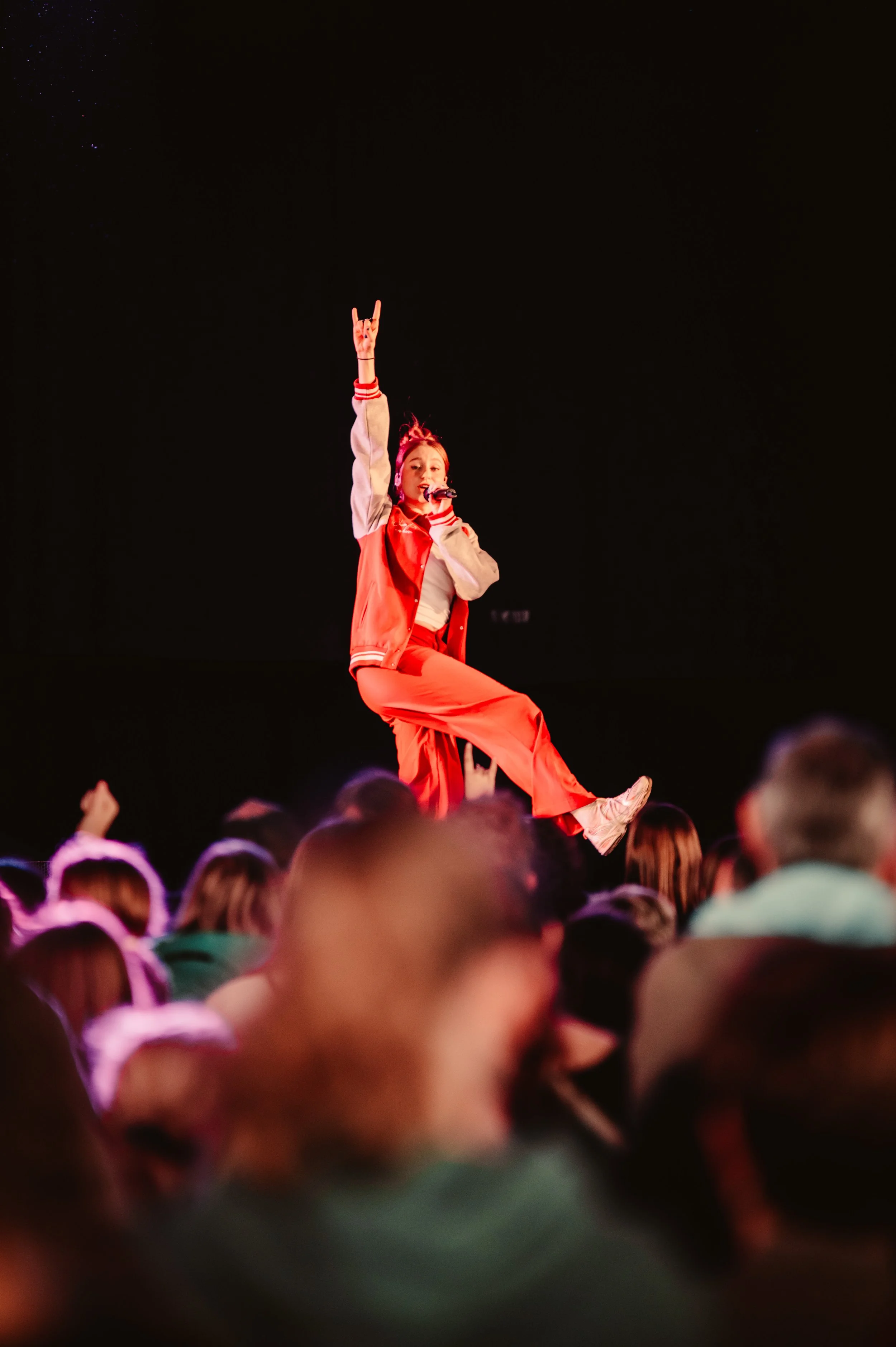 Performer in red outfit on stage with microphone and raised hand, crowd in foreground.