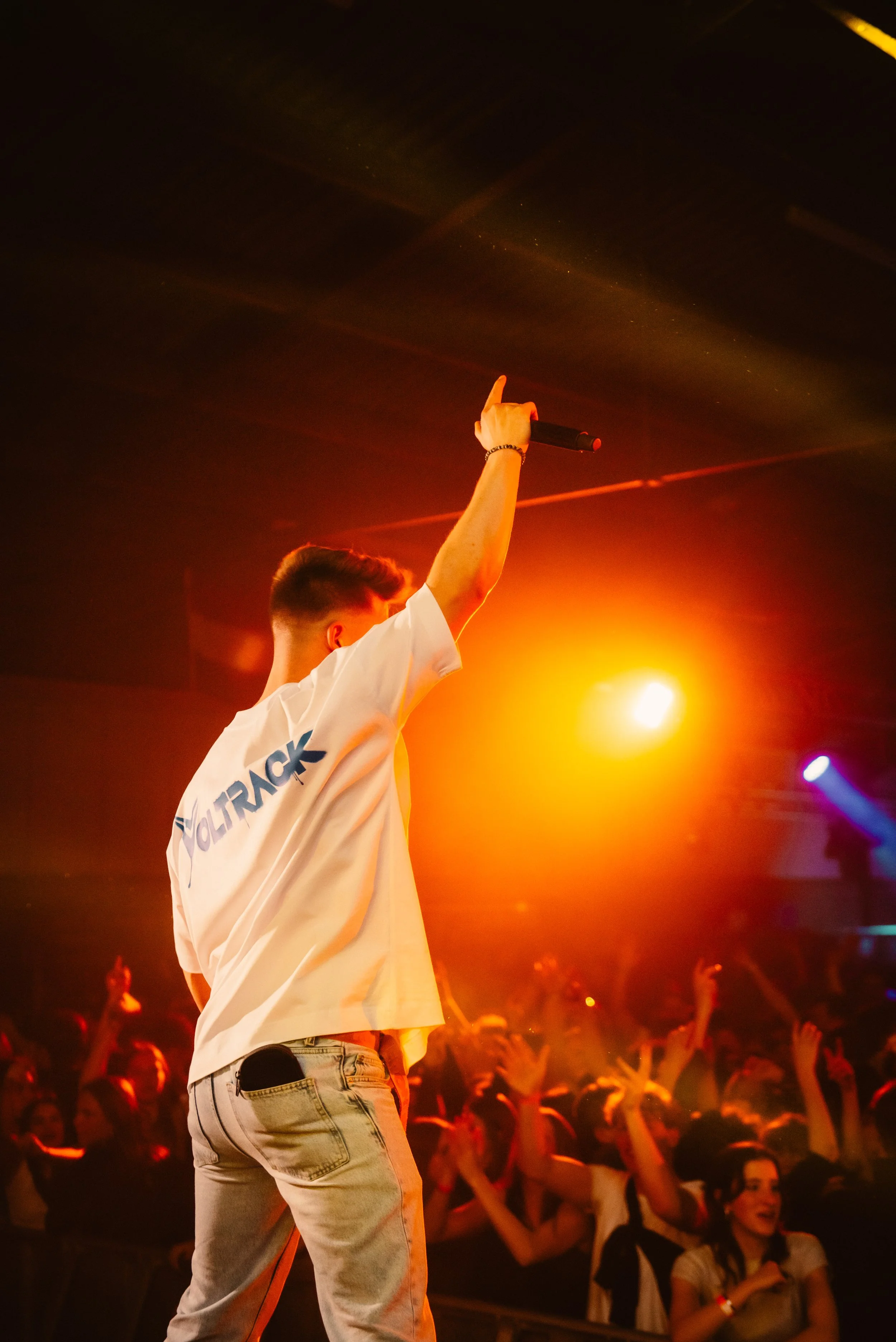 Performer on stage wearing a white shirt, holding a microphone, with one arm raised, facing a cheering crowd under bright stage lights.