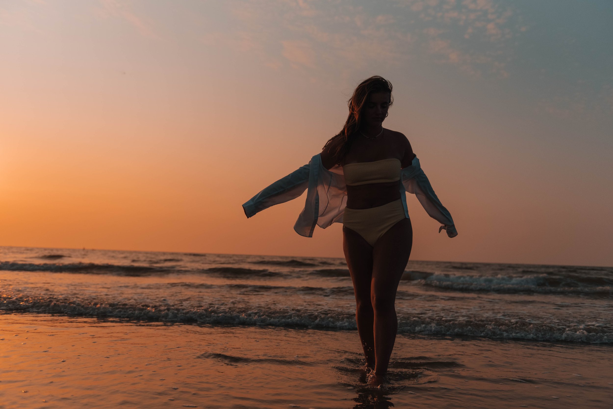 Silhouette of a woman walking in the ocean during sunset, with the sky displaying warm hues of orange and pink.