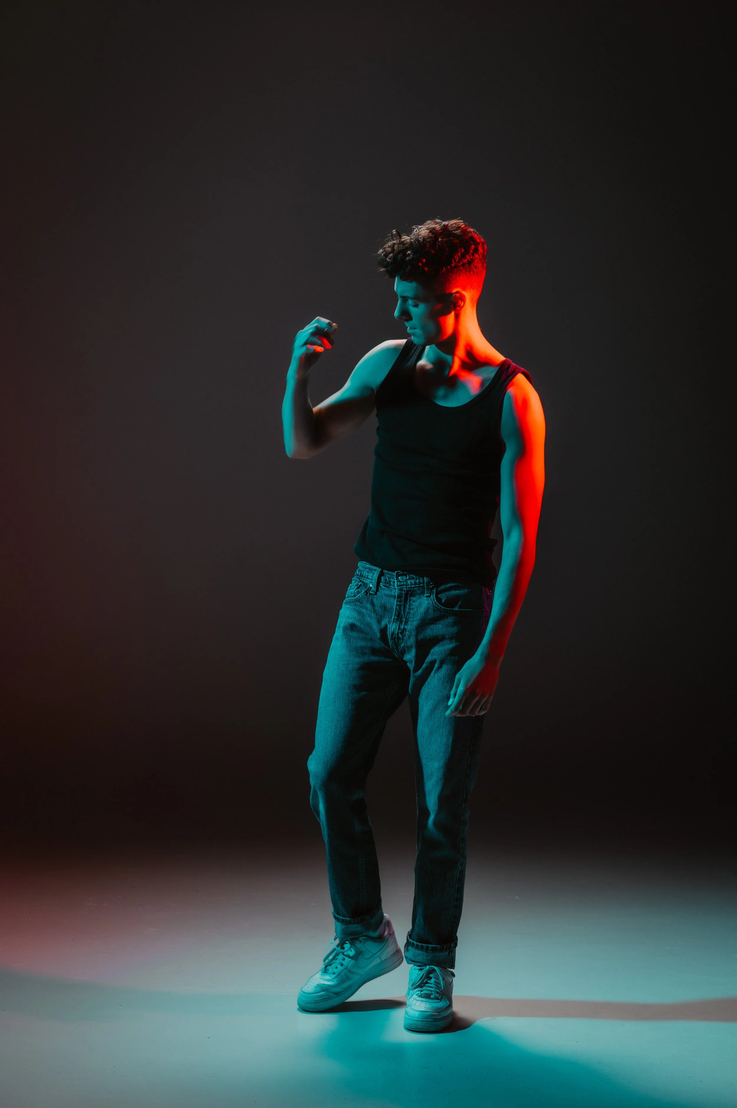 Person in dark room with artistic lighting, wearing a black tank top and jeans, looking at raised hand.