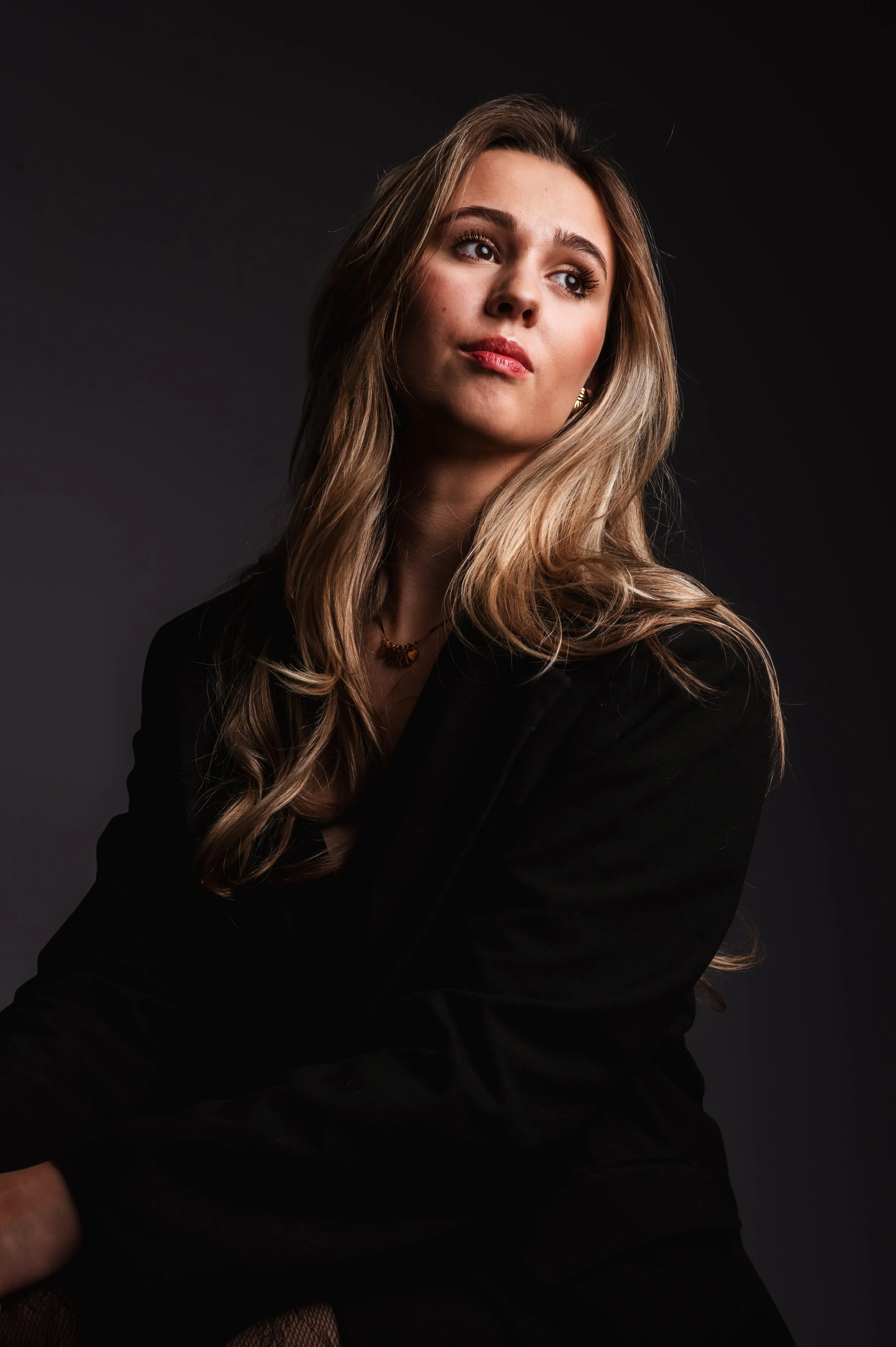 Portrait of a young woman with long wavy blonde hair, looking to the side, wearing a black jacket against a dark background.