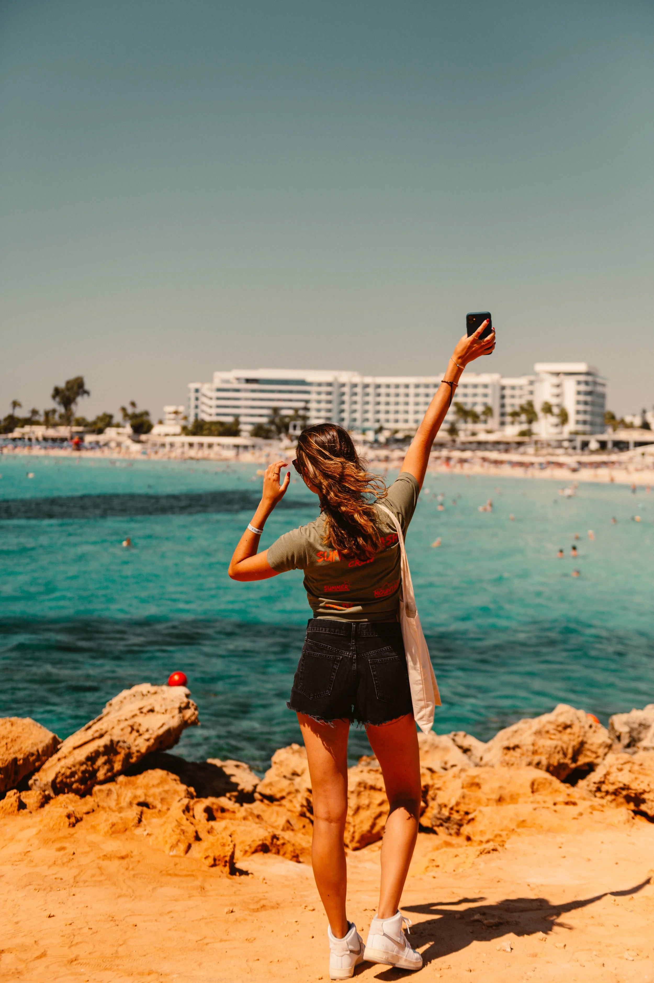 Woman taking selfie on rocky beach with hotel in background