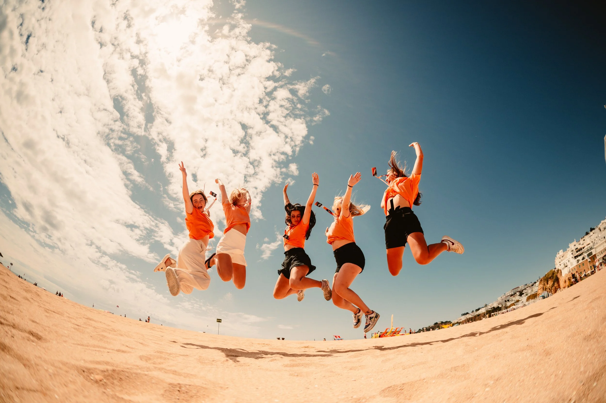 Group of people in orange shirts jumping on a beach under a clear sky.