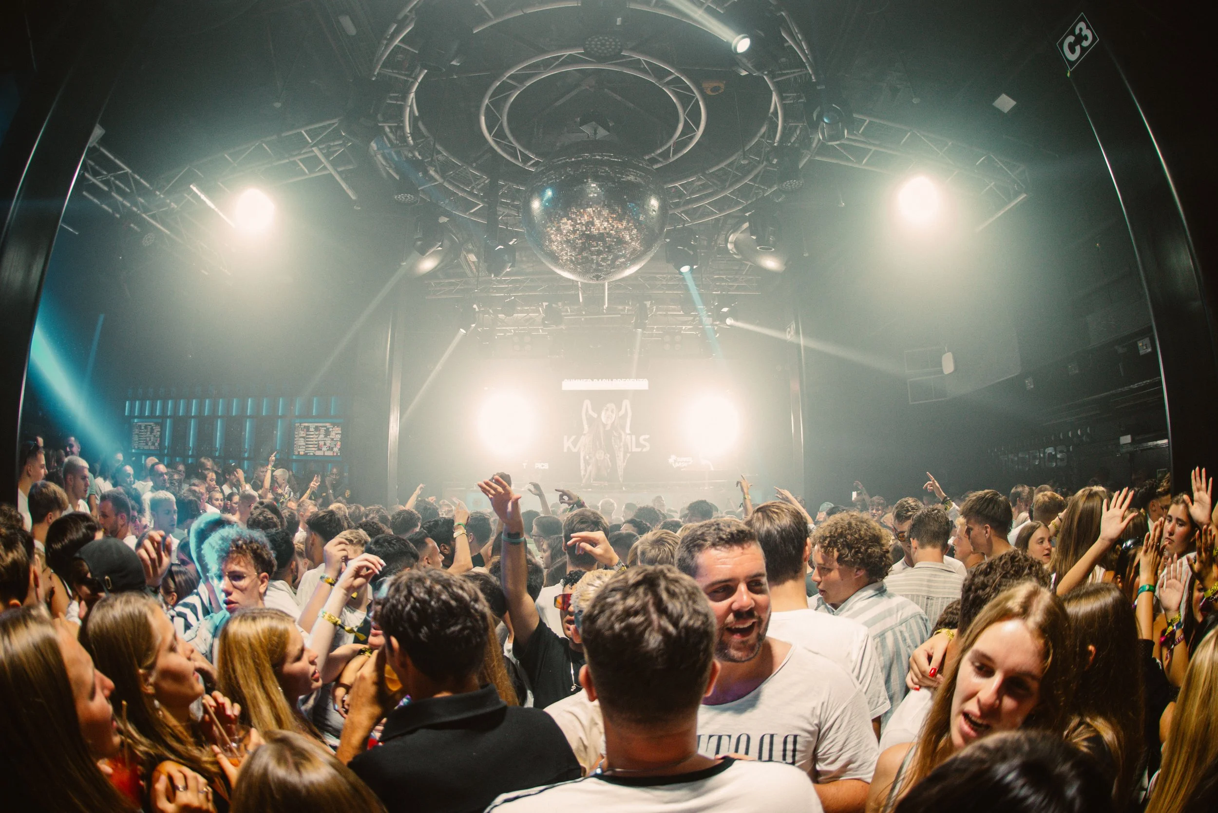 Crowd of people dancing and enjoying a concert at a nightclub with a brightly lit stage and disco ball.