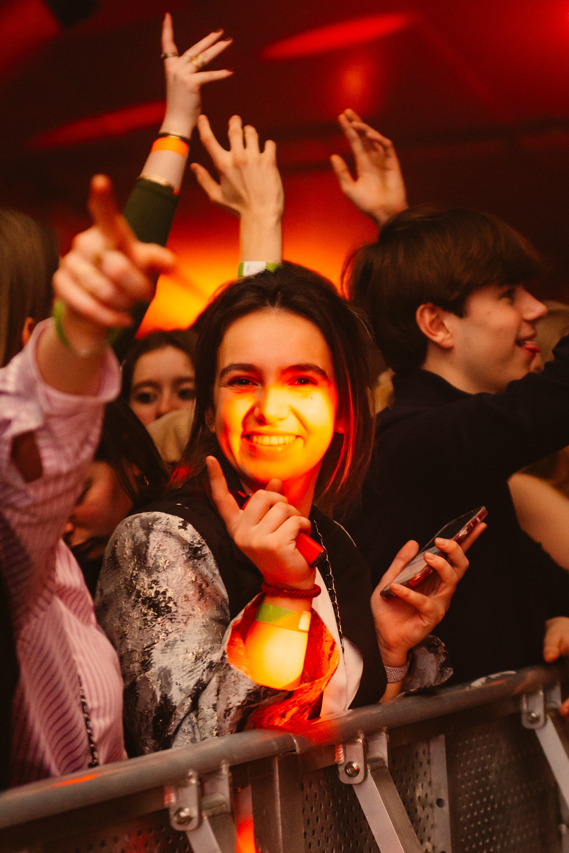 People enjoying a concert, smiling, holding a phone, with raised hands around them under red lighting.