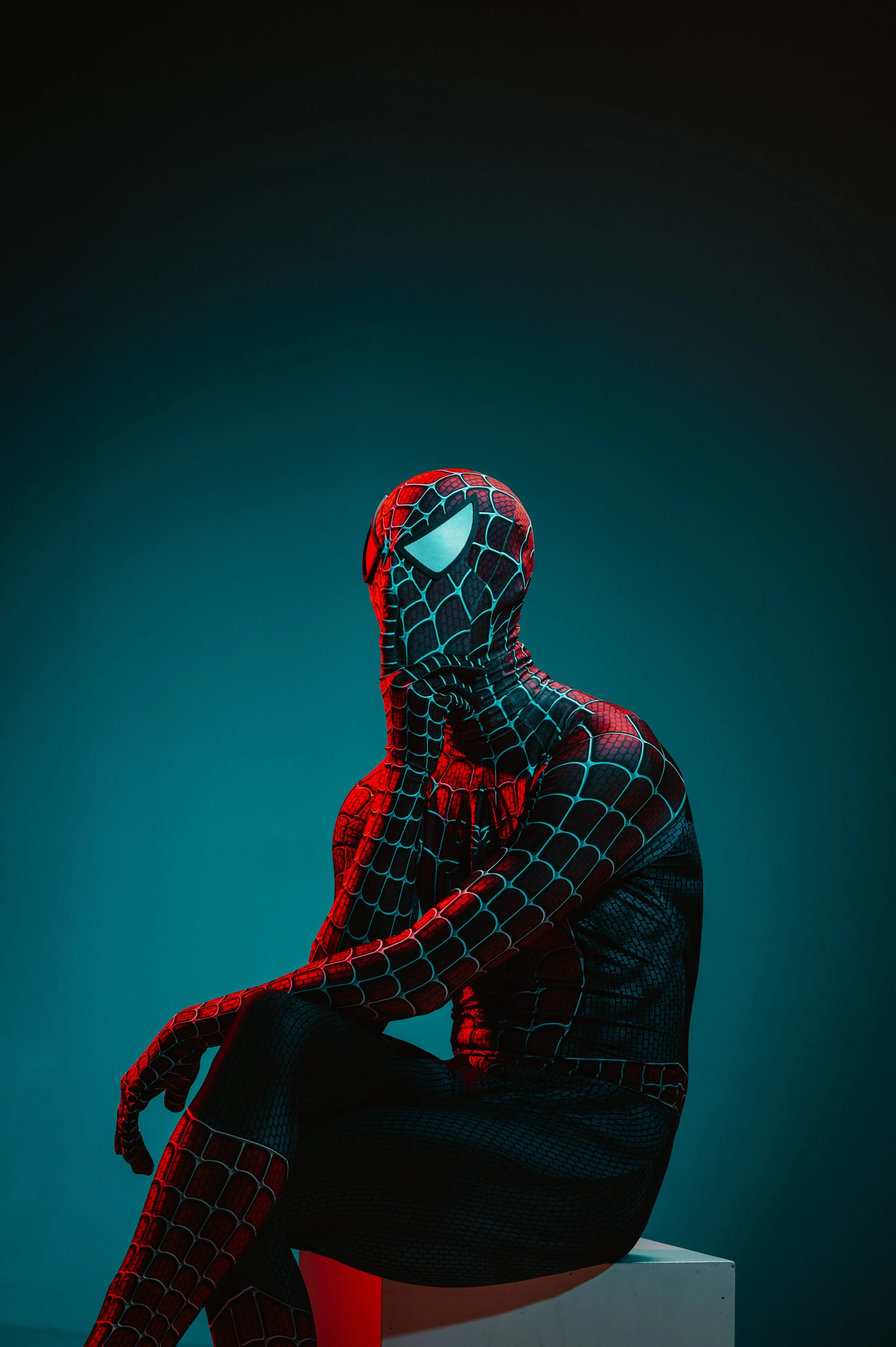 Person in a Spider-Man costume sitting on a cube, with a contemplative pose, under red and blue lighting.