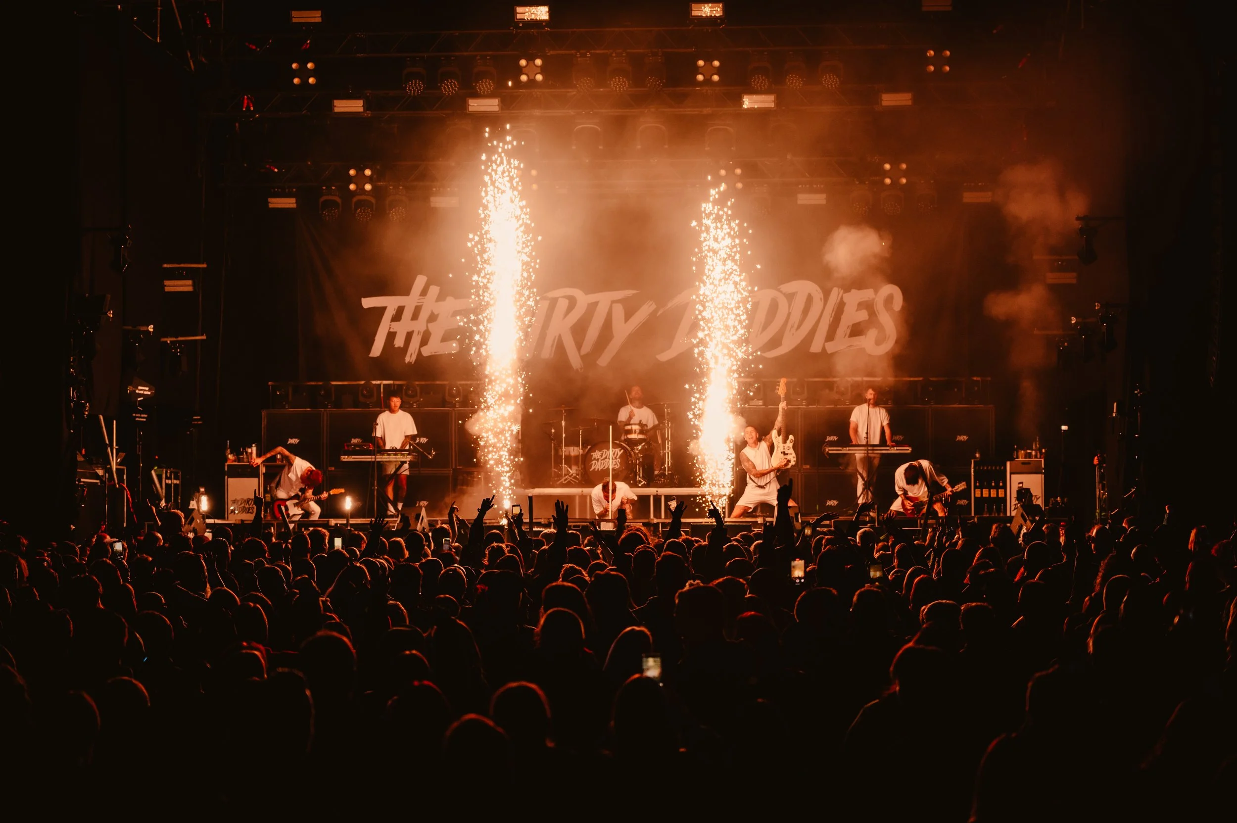 Concert stage with a band performing, pyrotechnics exploding, and a large crowd in the foreground.