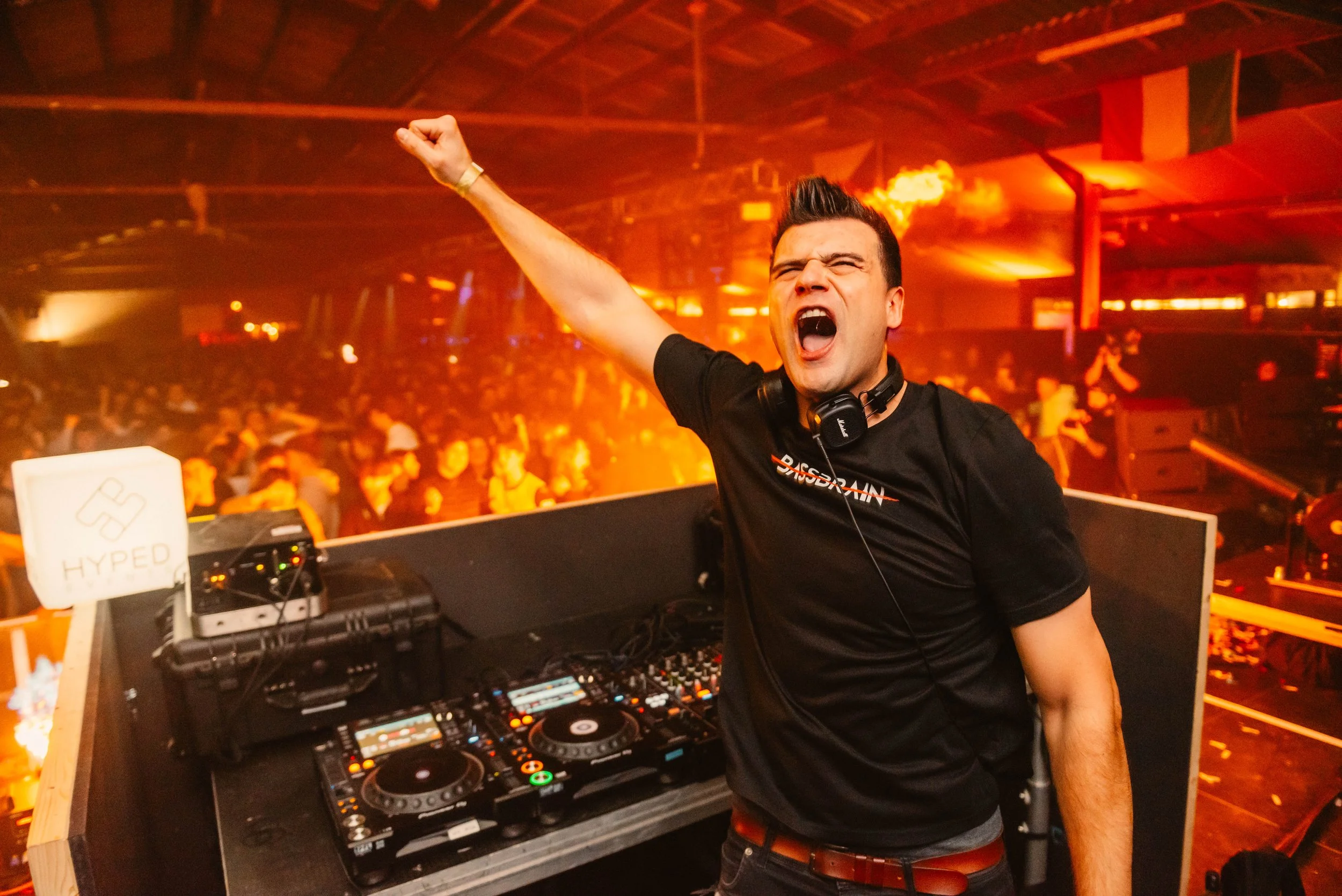 DJ performing live at a crowded nightclub, raising arm in excitement, with DJ equipment visible.