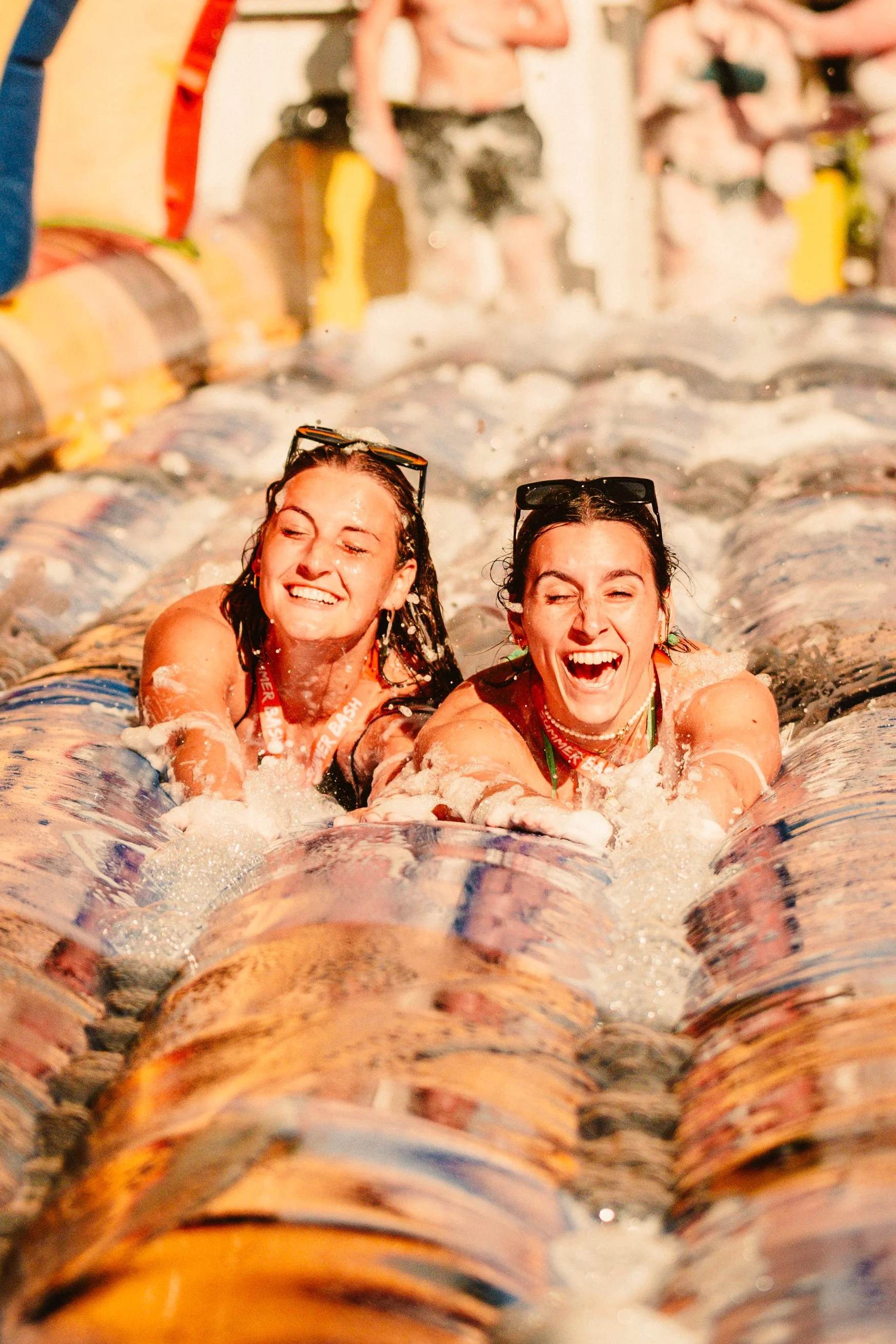 Two women enjoying a water slide, laughing and wearing swimsuits, with sunglasses on their heads. They are on a colorful inflatable slide in bright sunlight.