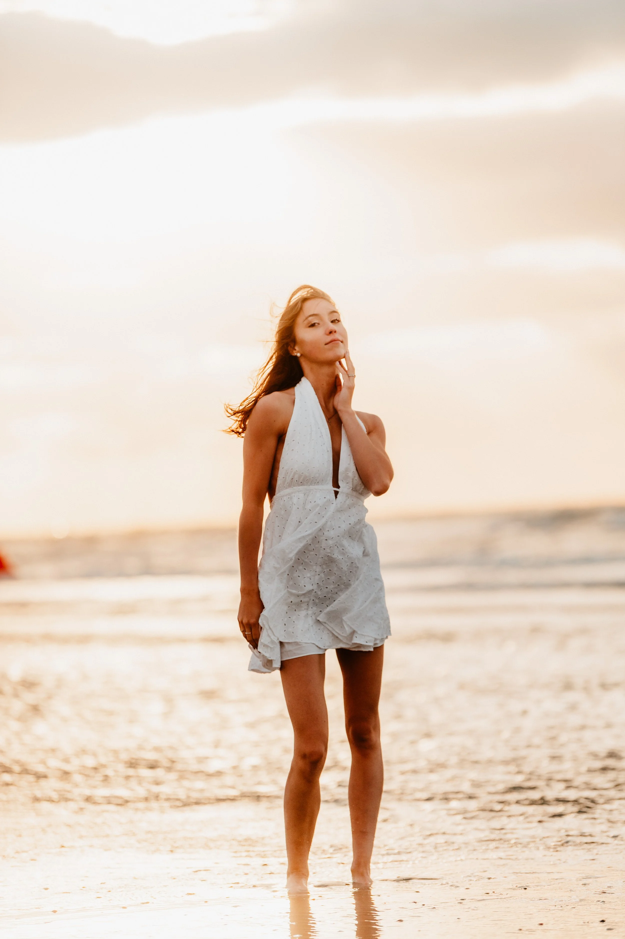 A woman in a white dress standing in shallow water at the beach during sunset.